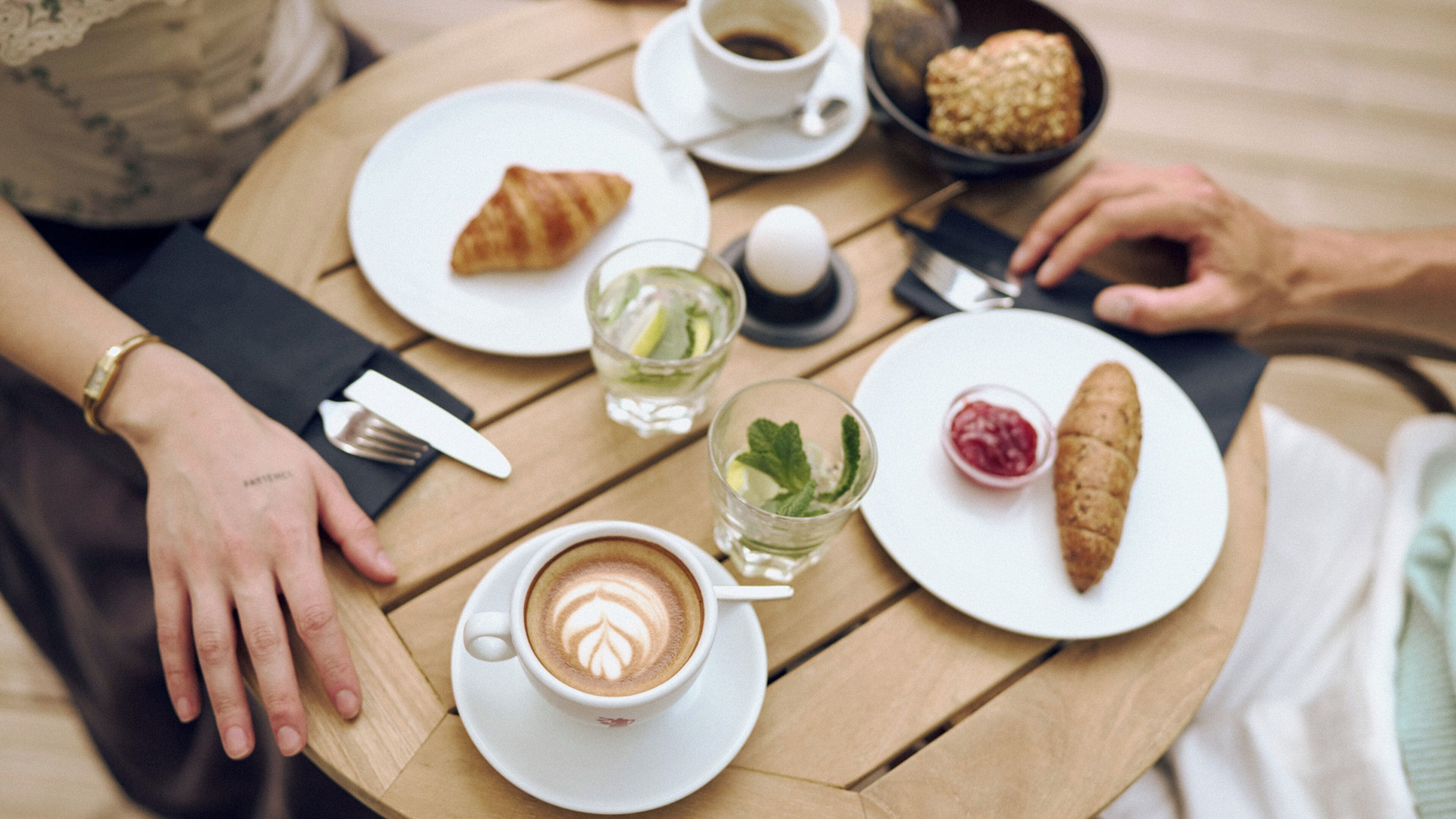 Une table ronde en bois, sur laquelle se trouvent une tasse de café, un croissant, un verre d’eau et un petit pot de confiture.