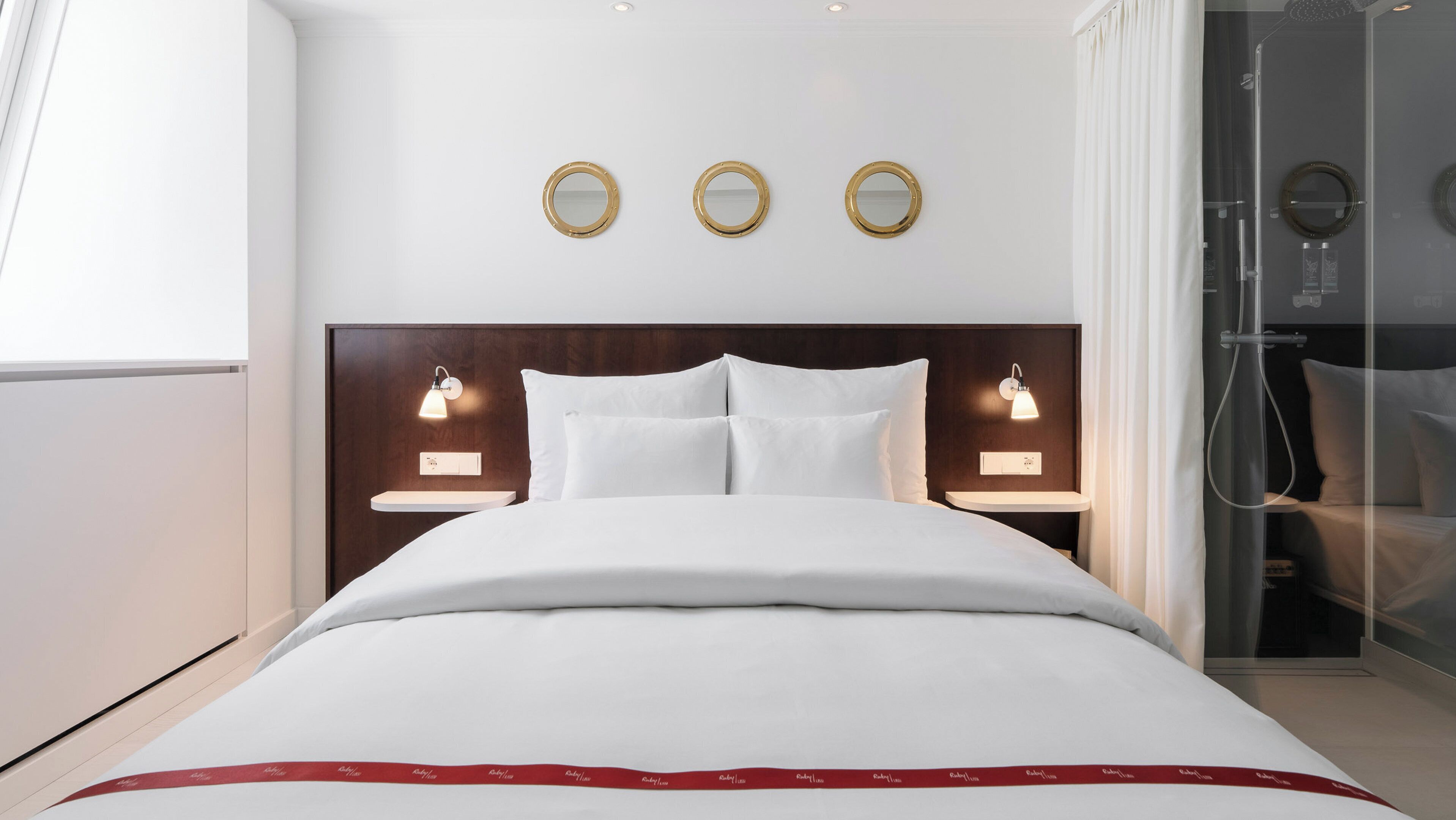A modern hotel room with a large bed, two bedside tables with lamps, a shower area, and three wall-mounted mirrors.
