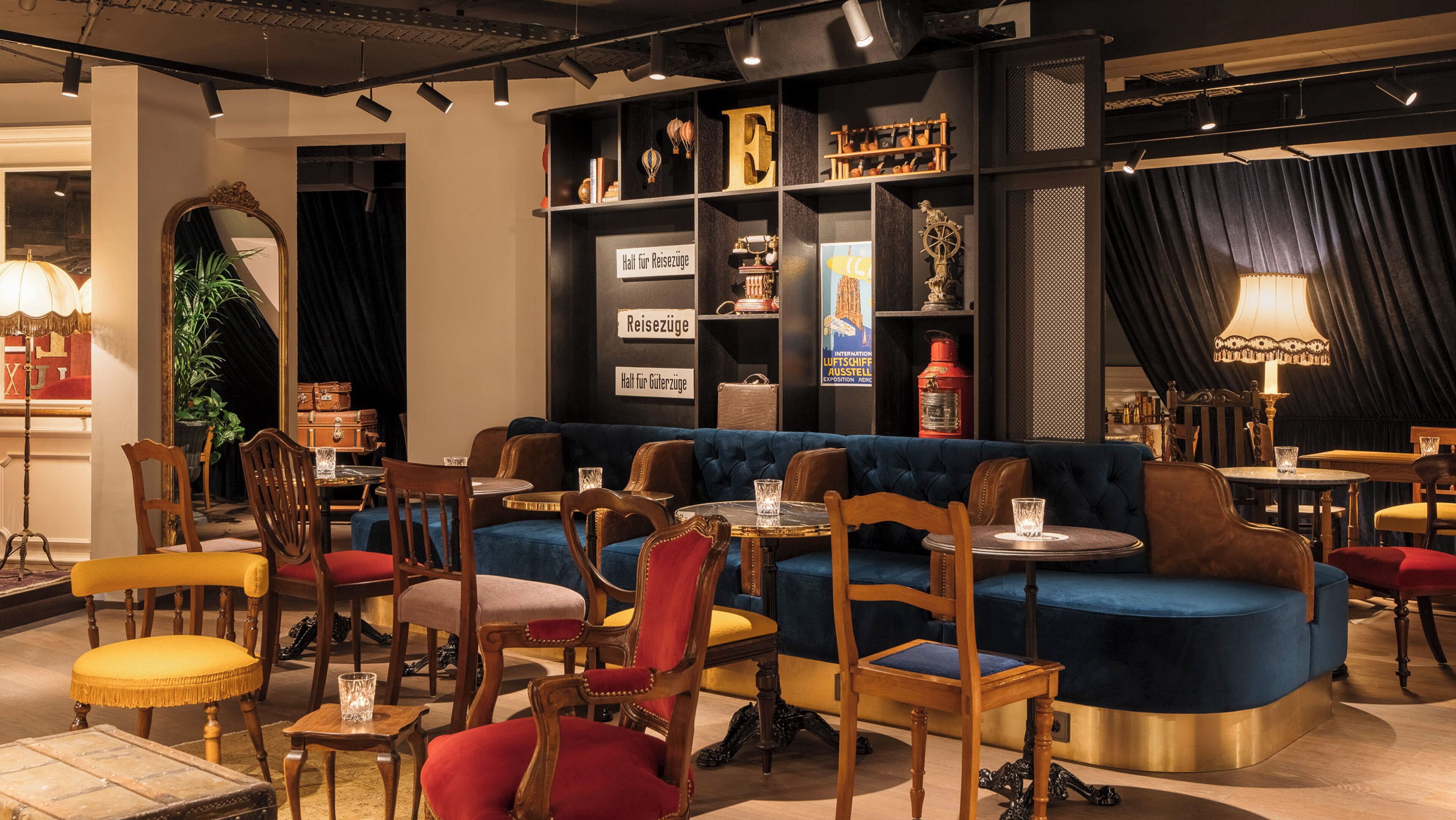 A cozy lounge area with a blue sofa, wooden chairs, and tables. There are suitcases and a sign indicating luggage storage.