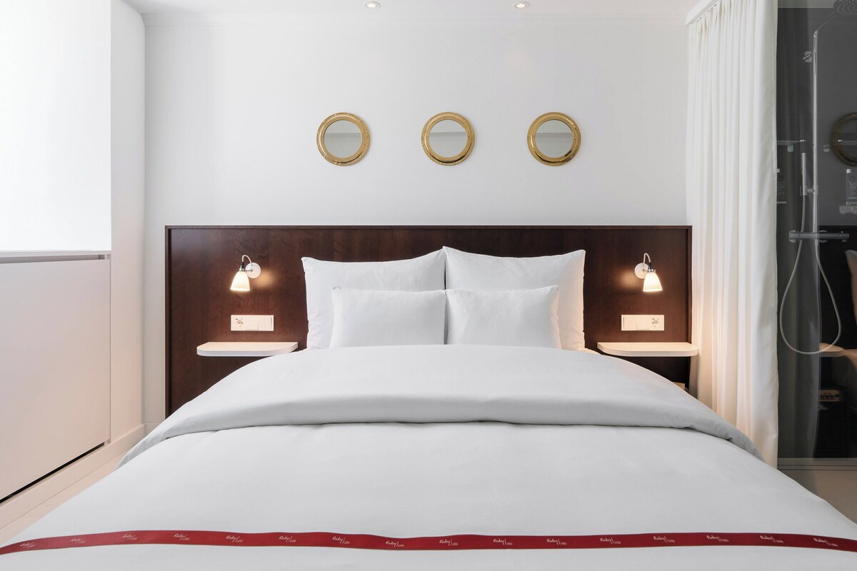 A modern hotel room with a large bed, two bedside tables with lamps, a shower area, and three wall-mounted mirrors.