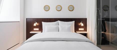 Una habitación de hotel moderna con una cama grande, dos mesitas de noche con lámparas, zona de ducha y tres espejos fijos a la pared.