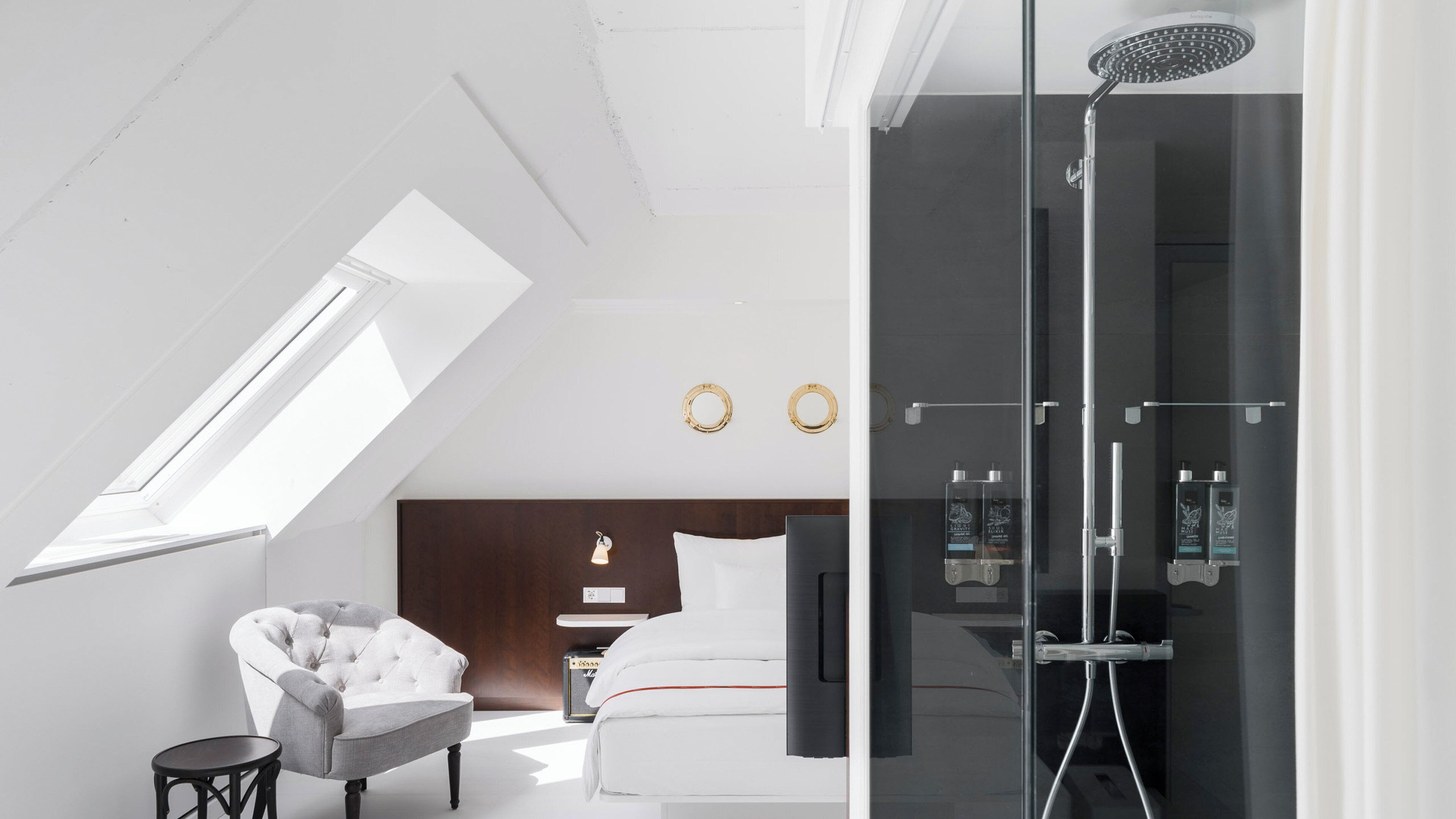 A modern hotel room with a skylight, a glass-enclosed shower, a comfortable armchair, and a neatly made bed.