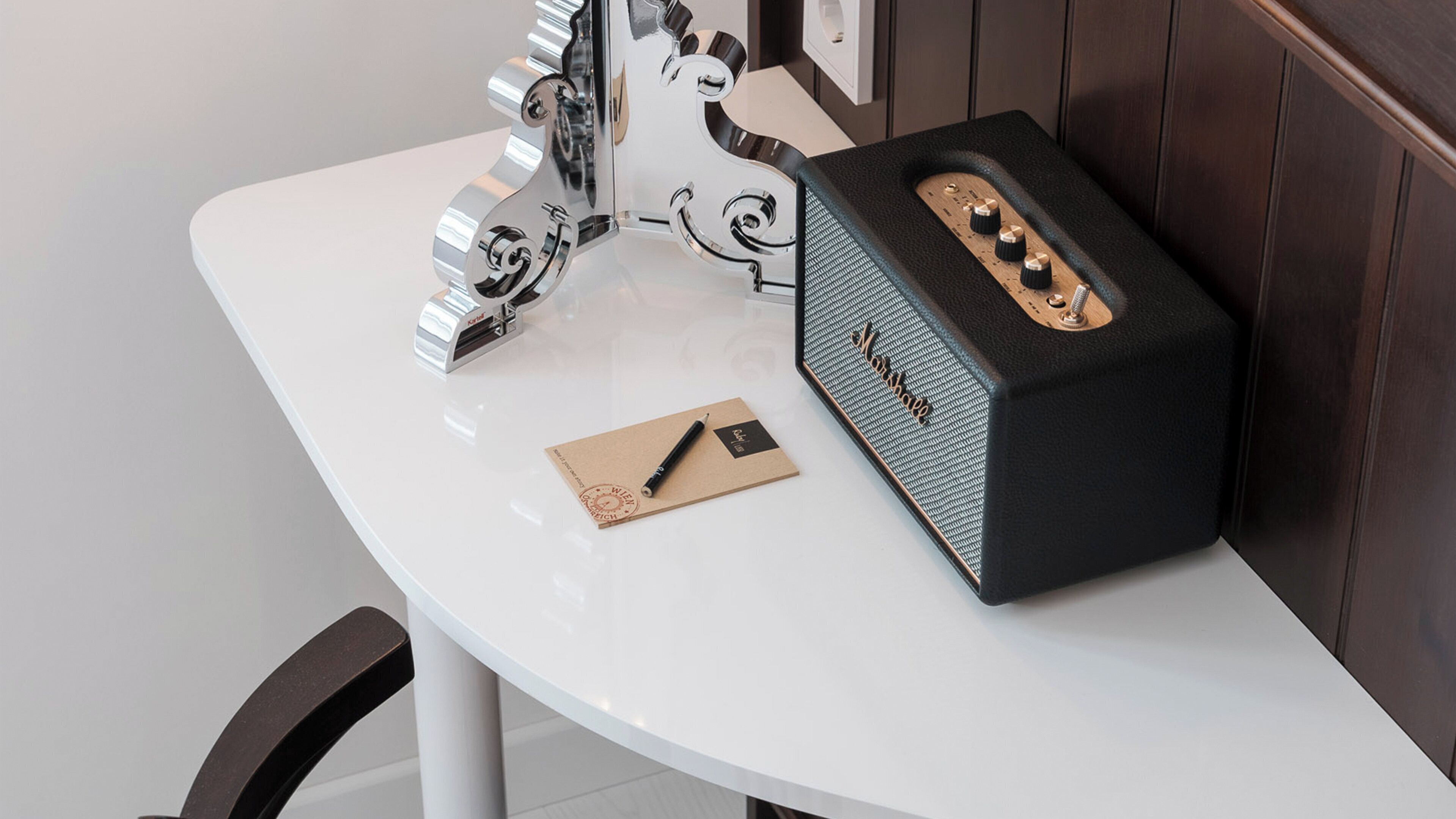 Une table blanche avec un haut-parleur noir, un objet argenté et une enveloppe marron avec un stylo.