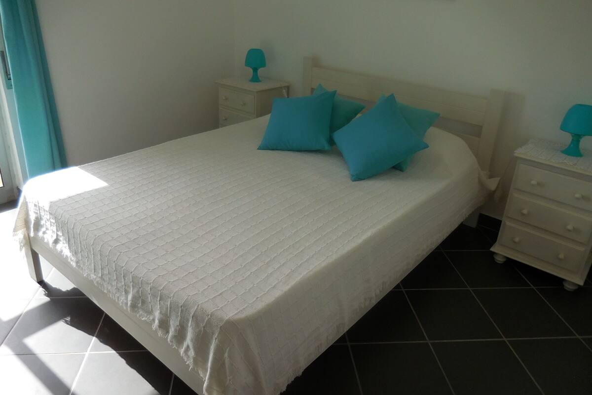 A neatly made bed with a white bedspread and blue pillows, a small bedside table with a lamp, and a nightstand with a lamp.