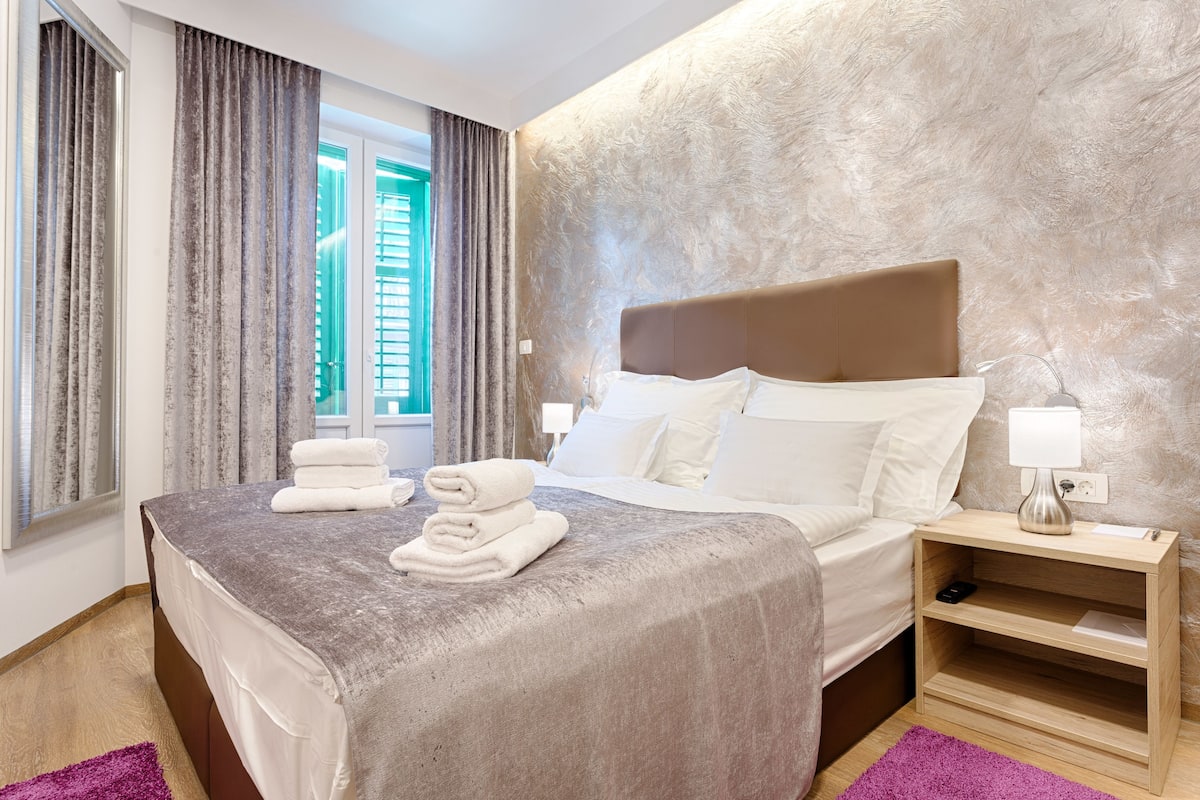 A hotel room with a large bed, a nightstand, a window with curtains, and a textured wall.