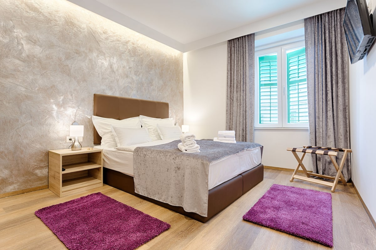 A modern hotel room with a large bed, a wooden nightstand, a small table, a window with curtains, and a wall-mounted TV.