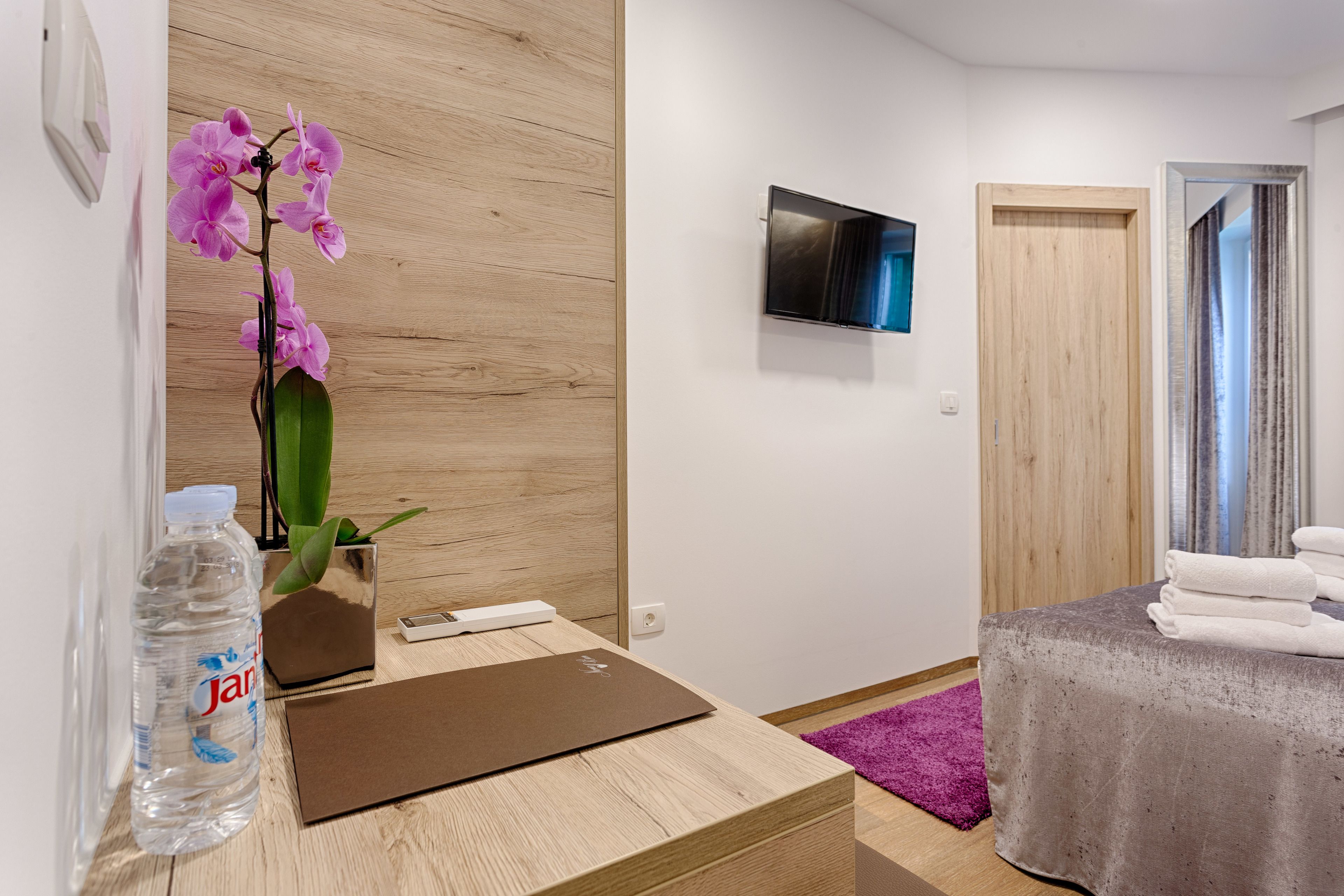 A hotel room with a wooden desk, a laptop, a water bottle, a potted orchid, a TV, a bed with towels, and a wooden door.