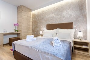 Una habitación de hotel moderna con una cama grande, cabecera de madera, mesitas de noche con lámparas, un pequeño escritorio con un florero y una alfombra rosa.