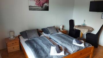 A bedroom with a wooden bed, grey bedding, a small bedside table with a lamp, a chair, a round table with a bottle, and a TV on the wall.