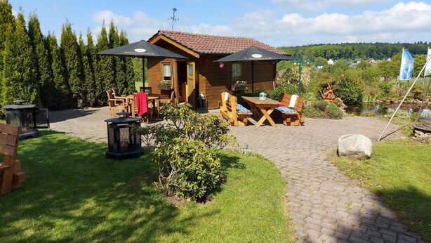 Eine Holzhütte mit einer Terrasse, Sitzgelegenheiten im Freien und Blick auf einen See und einen Wald.