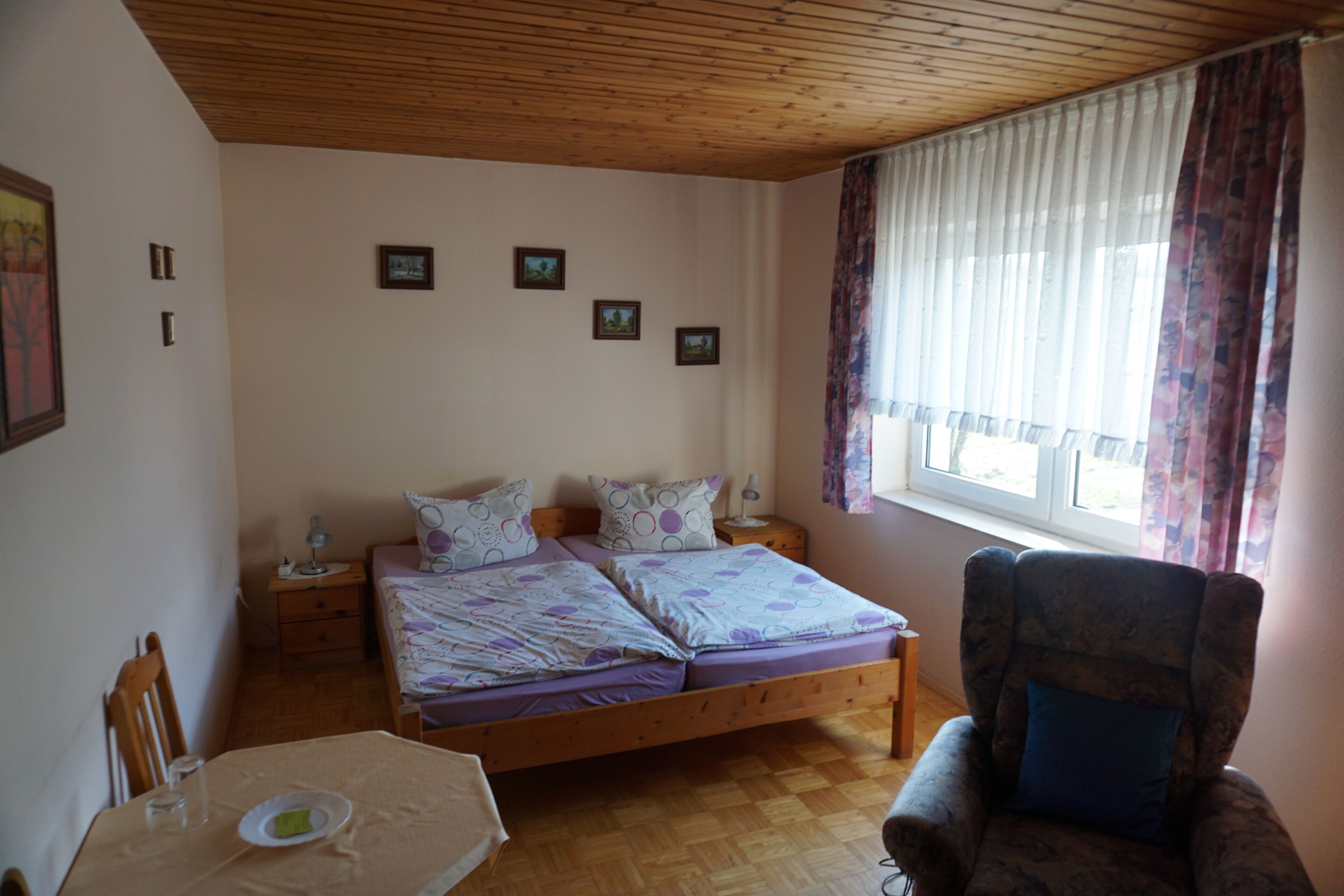 Une chambre avec un plafond en bois, un lit double avec une literie violette, une chaise, une table avec une assiette et une fenêtre avec des rideaux.
