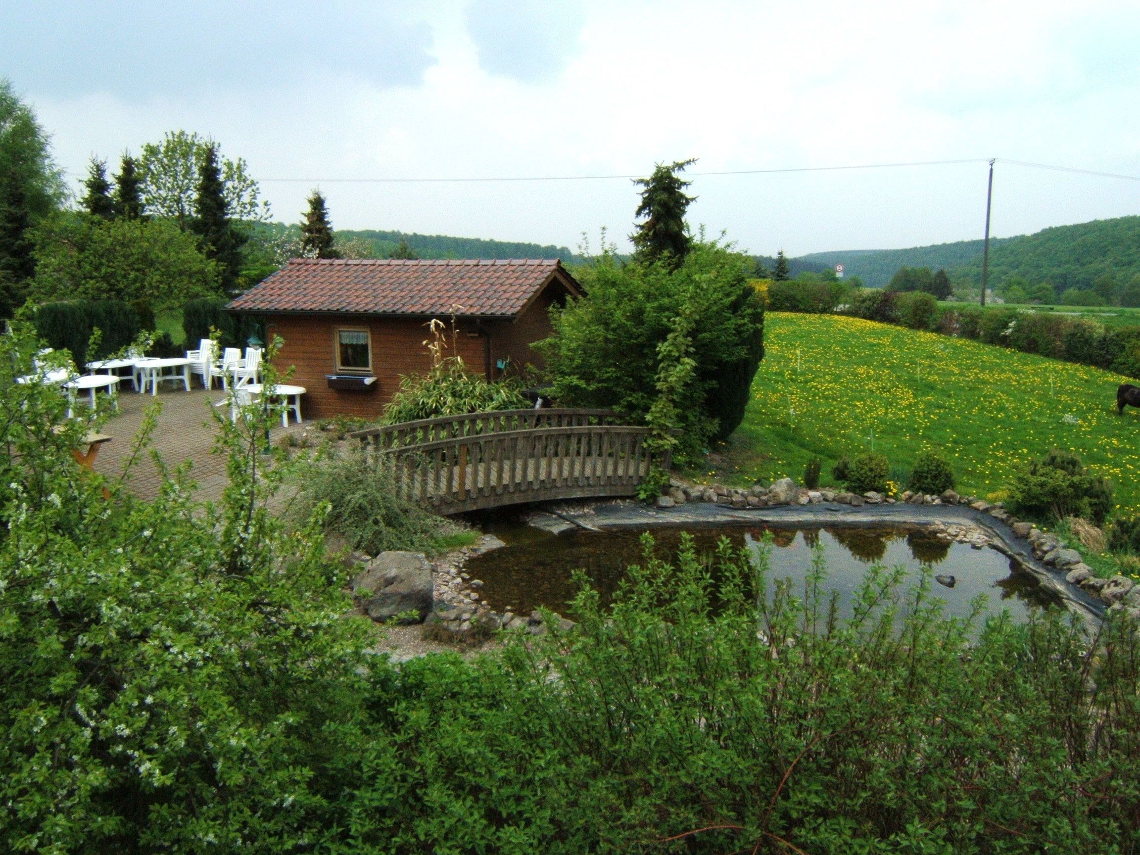 Una casa rústica con un pequeño puente sobre un estanque, rodeada de follaje y un campo de flores amarillas.