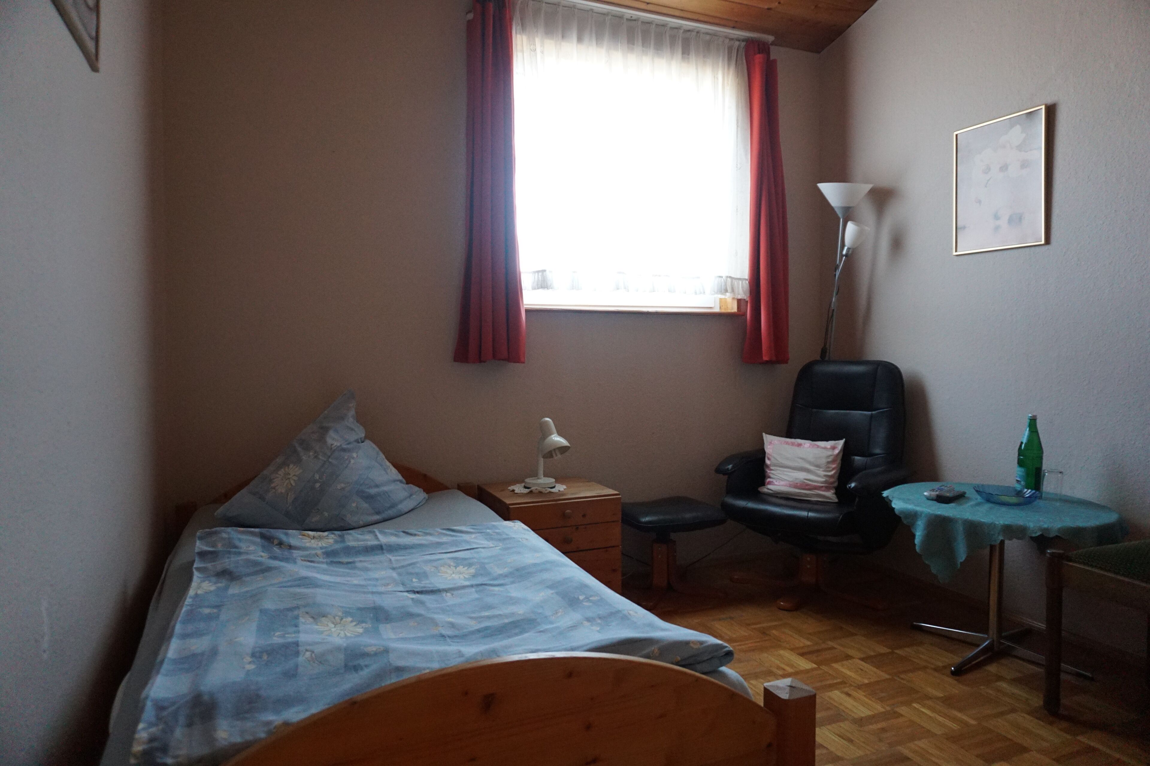 Ein Schlafzimmer mit einem Bett, einem Stuhl, einem Schreibtisch, einer Lampe und einem Fenster mit Vorhängen.