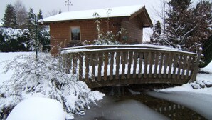 Eine Holzhütte mit schneebedecktem Dach und einer Holzbrücke über einem kleinen Bach.