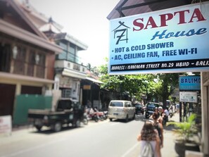 A street view with a sign for 'SAPTA House' advertising amenities like a hot & cold shower, AC, ceiling fan, and free Wi-Fi. - Sapta House (Ubud)