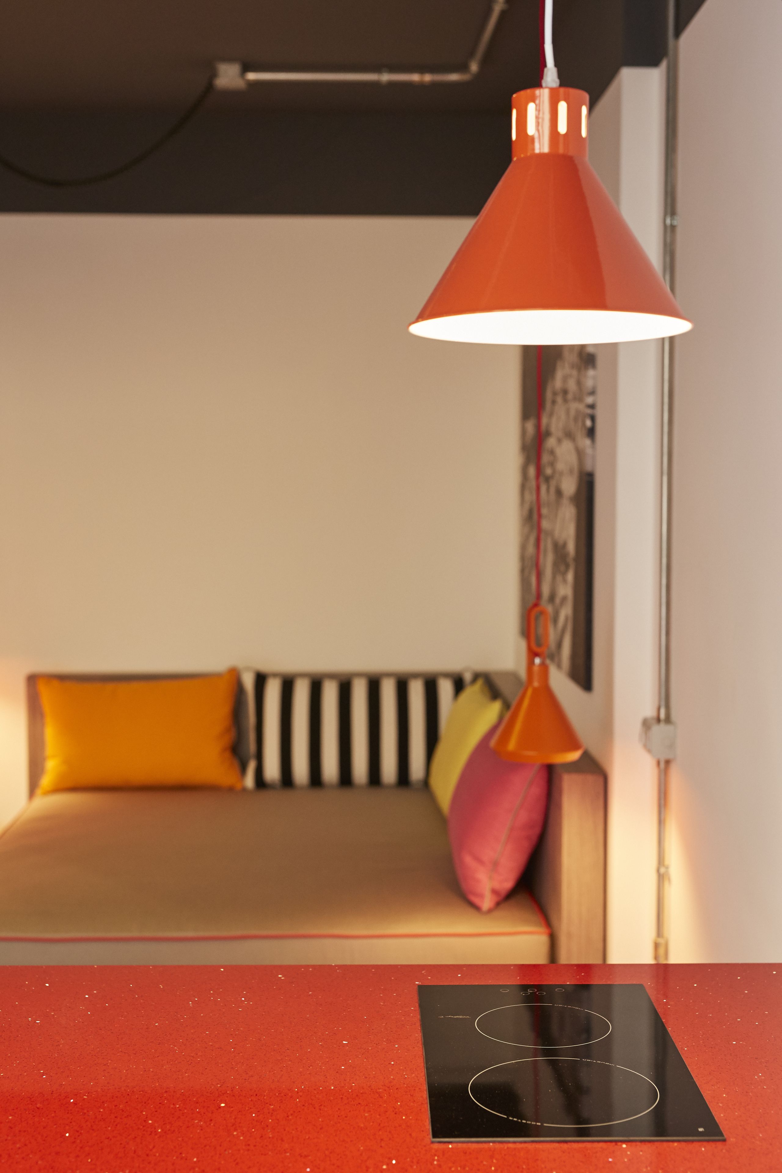 A modern living room with a sofa, a red table with a black surface, and a hanging orange pendant light.