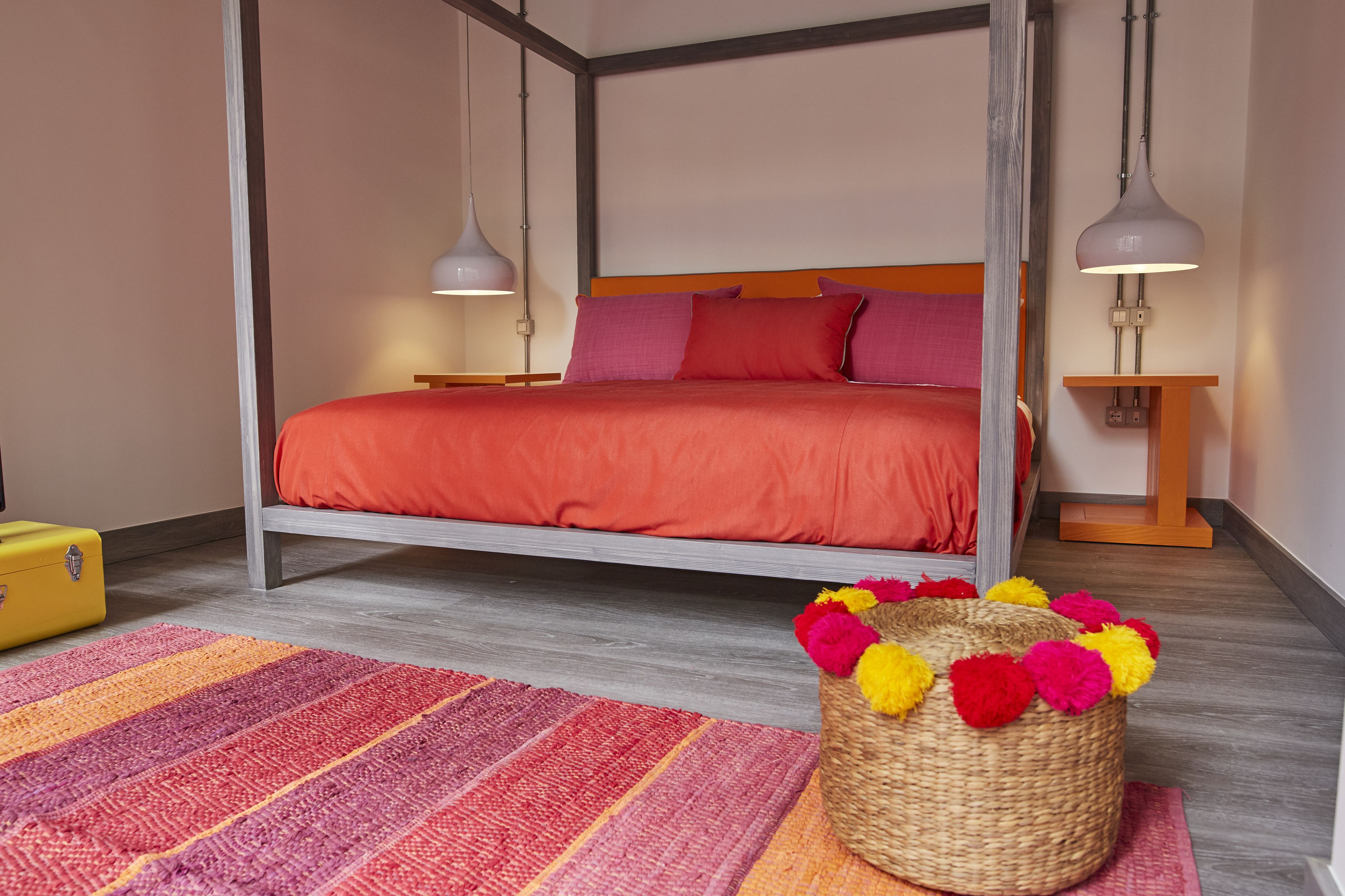 A four-poster bed with a red bedspread, two bedside lamps, a wicker basket with colorful pom-poms, and a striped rug.
