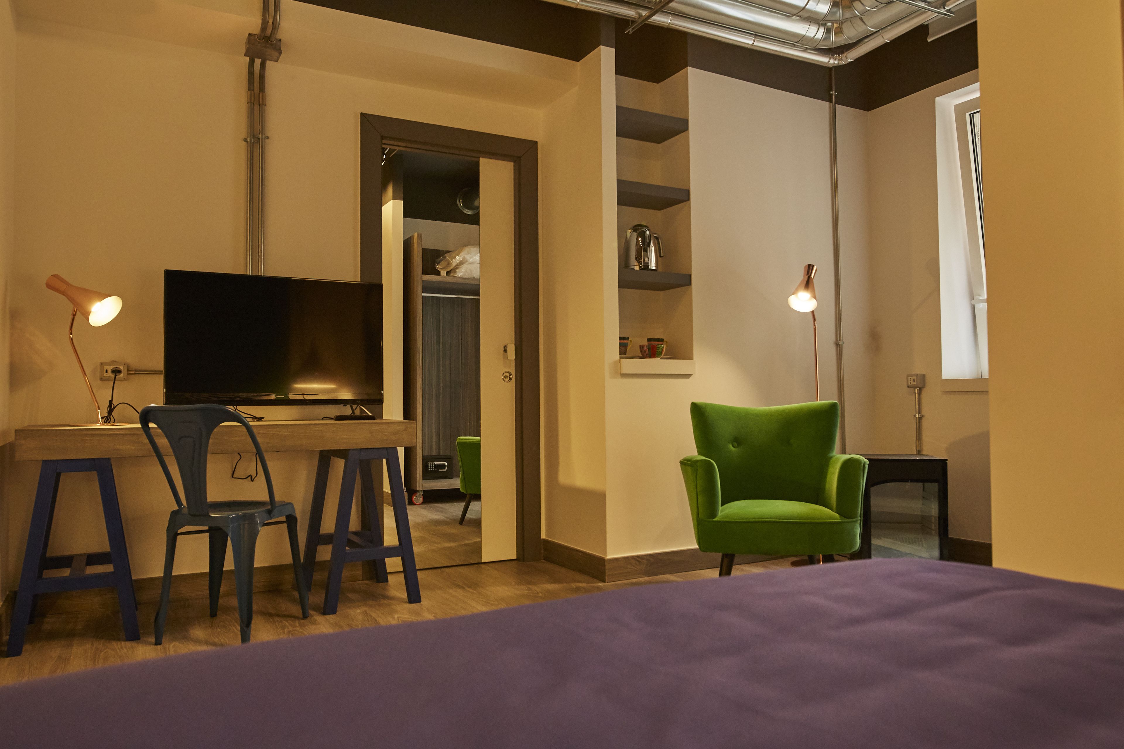 A room with a bed, a desk with a computer, two chairs, a green armchair, a television, and a shelving unit with items.