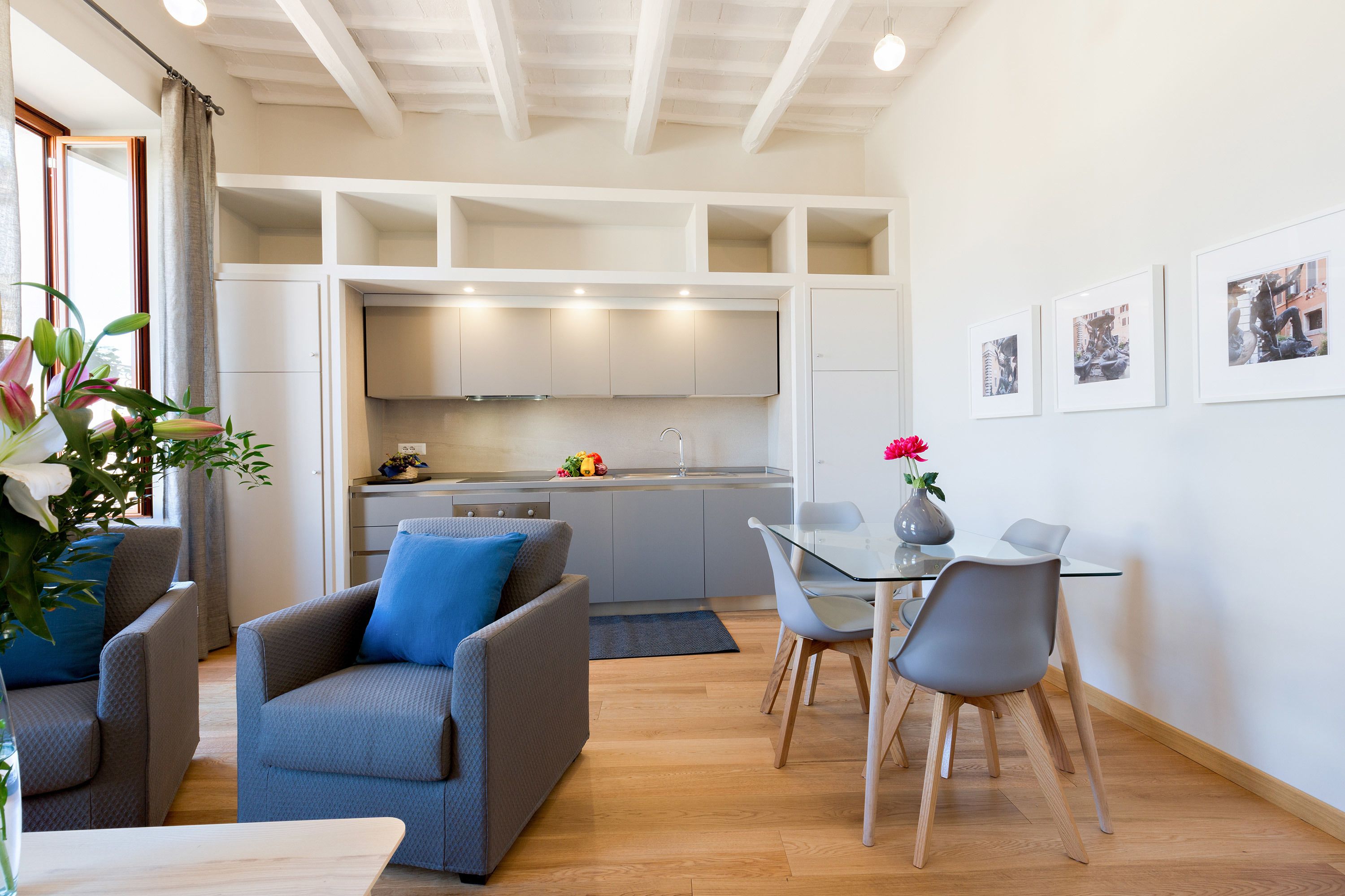Una cucina moderna con mobili bianchi, un'area pranzo con tavolo in vetro e sedie grigie, e un divano grigio con cuscini blu.