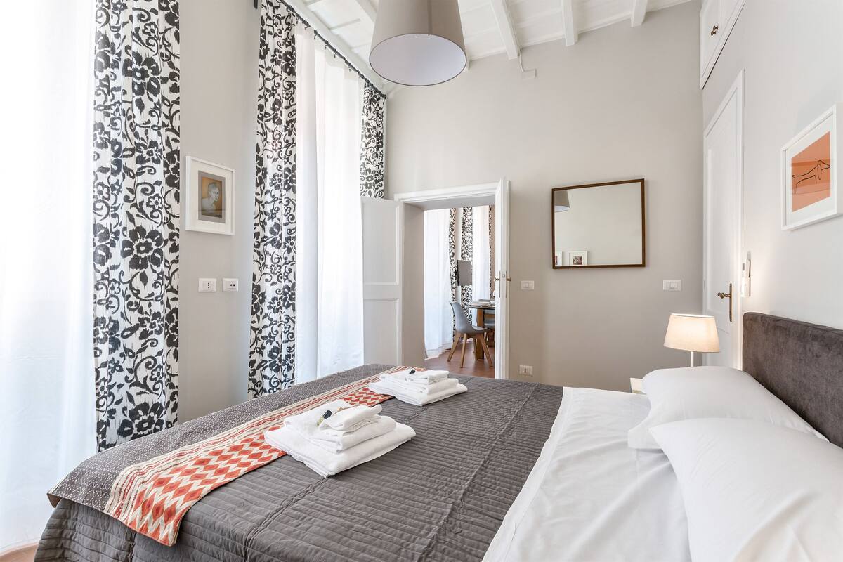 A neatly made bed with a grey quilt, white pillows, and folded towels. There is a mirror on the wall and a lamp beside the bed.
