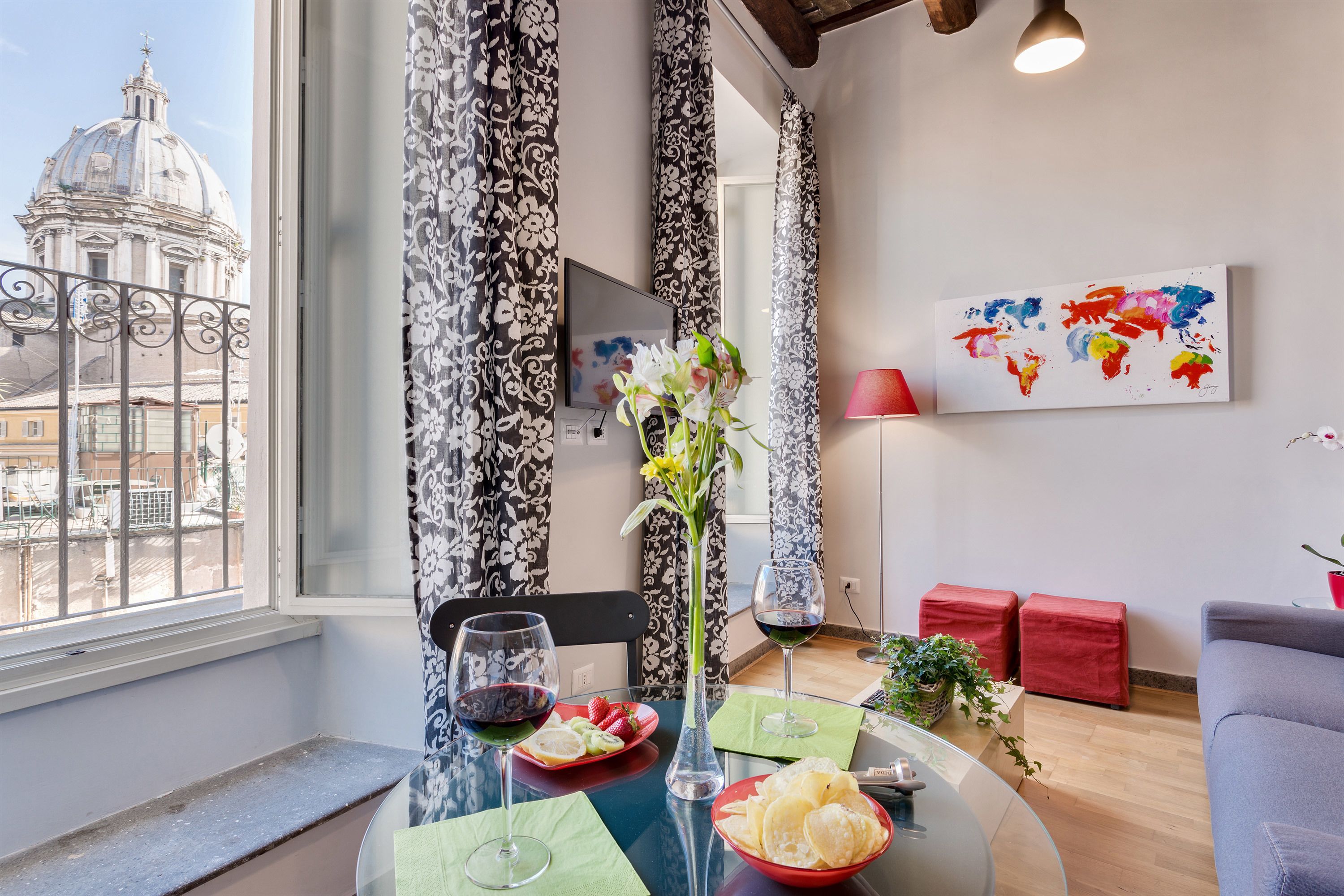 A modern living room with a dining area, featuring a large window with a view of a historic building, a world map artwork, and a glass table with a vase of flowers.
