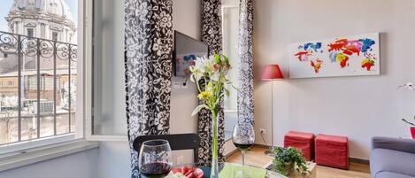 A modern living room with a dining area, featuring a large window with a view of a historic building, a world map artwork, and a glass table with a vase of flowers.