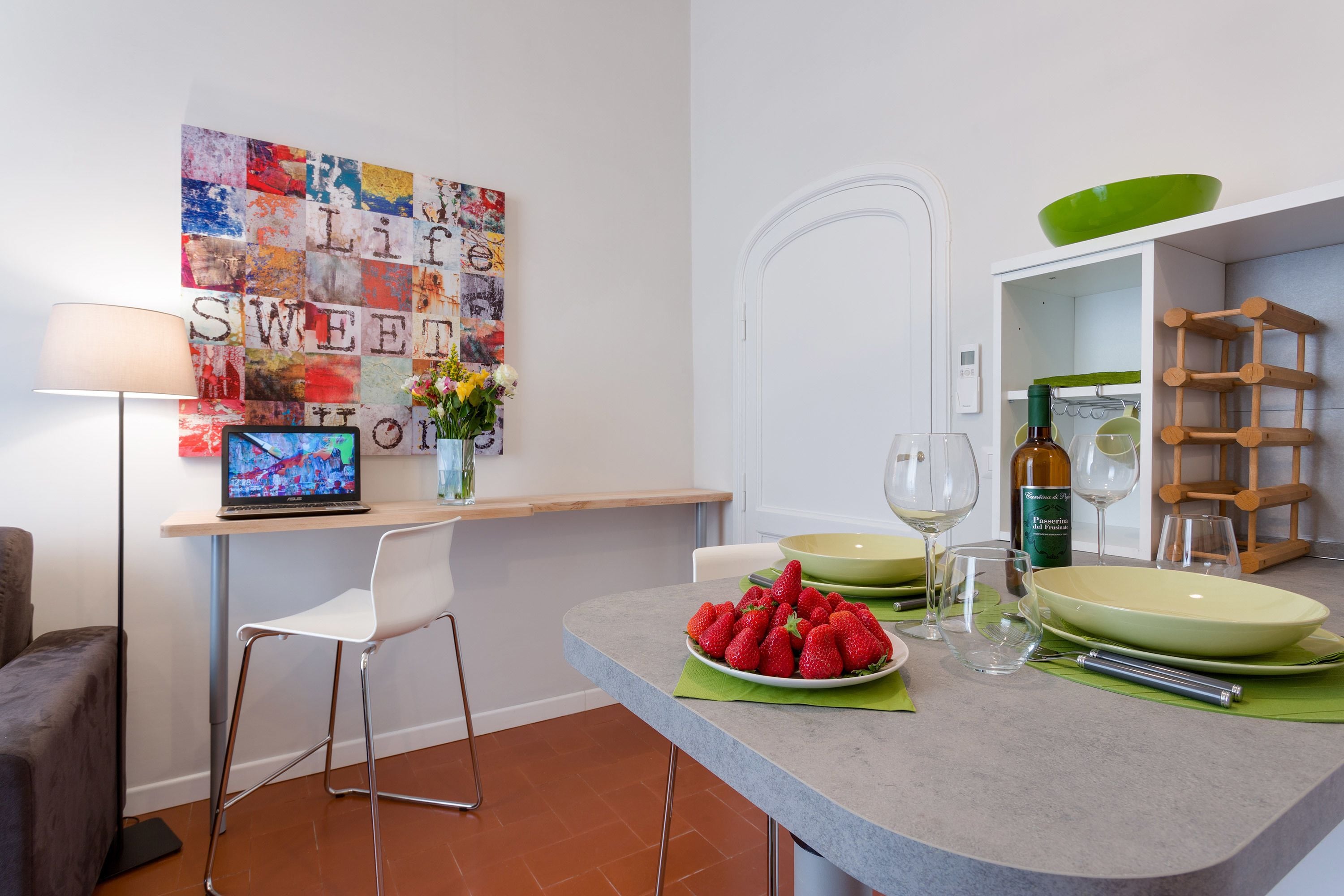 A modern kitchen with a laptop on a counter, a bowl of strawberries, wine glasses, and a green bowl on the shelf.
