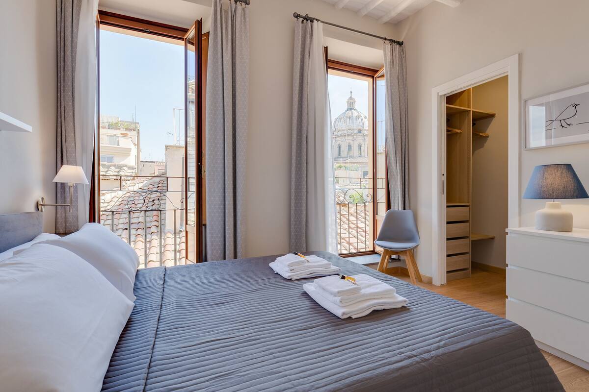 A hotel room with a bed, two towels, a chair, a desk, and a view of a cityscape through large windows.