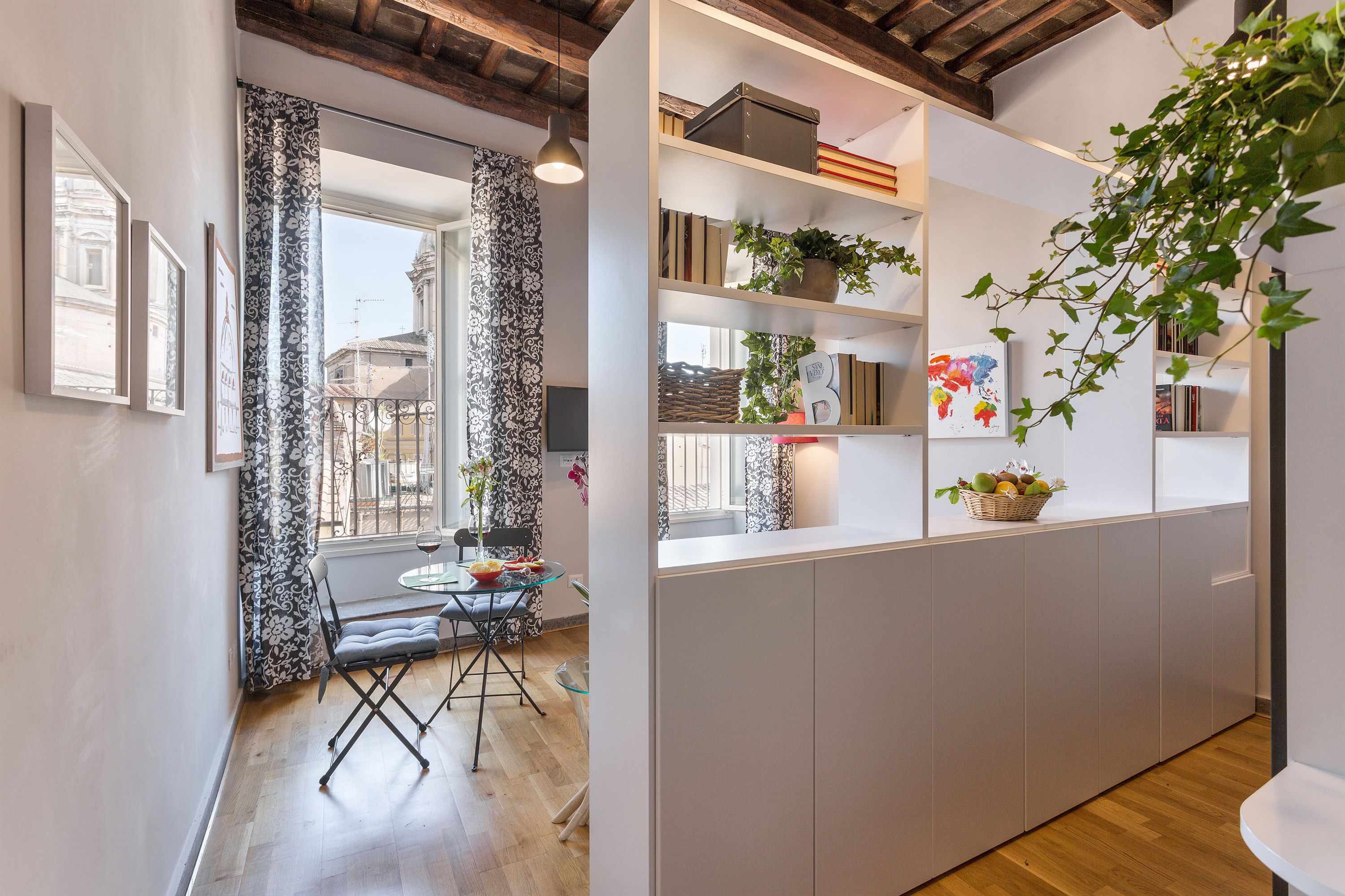 Cozinha moderna com vigas de madeira, área de jantar com mesa e cadeiras, e uma estante com plantas e livros.