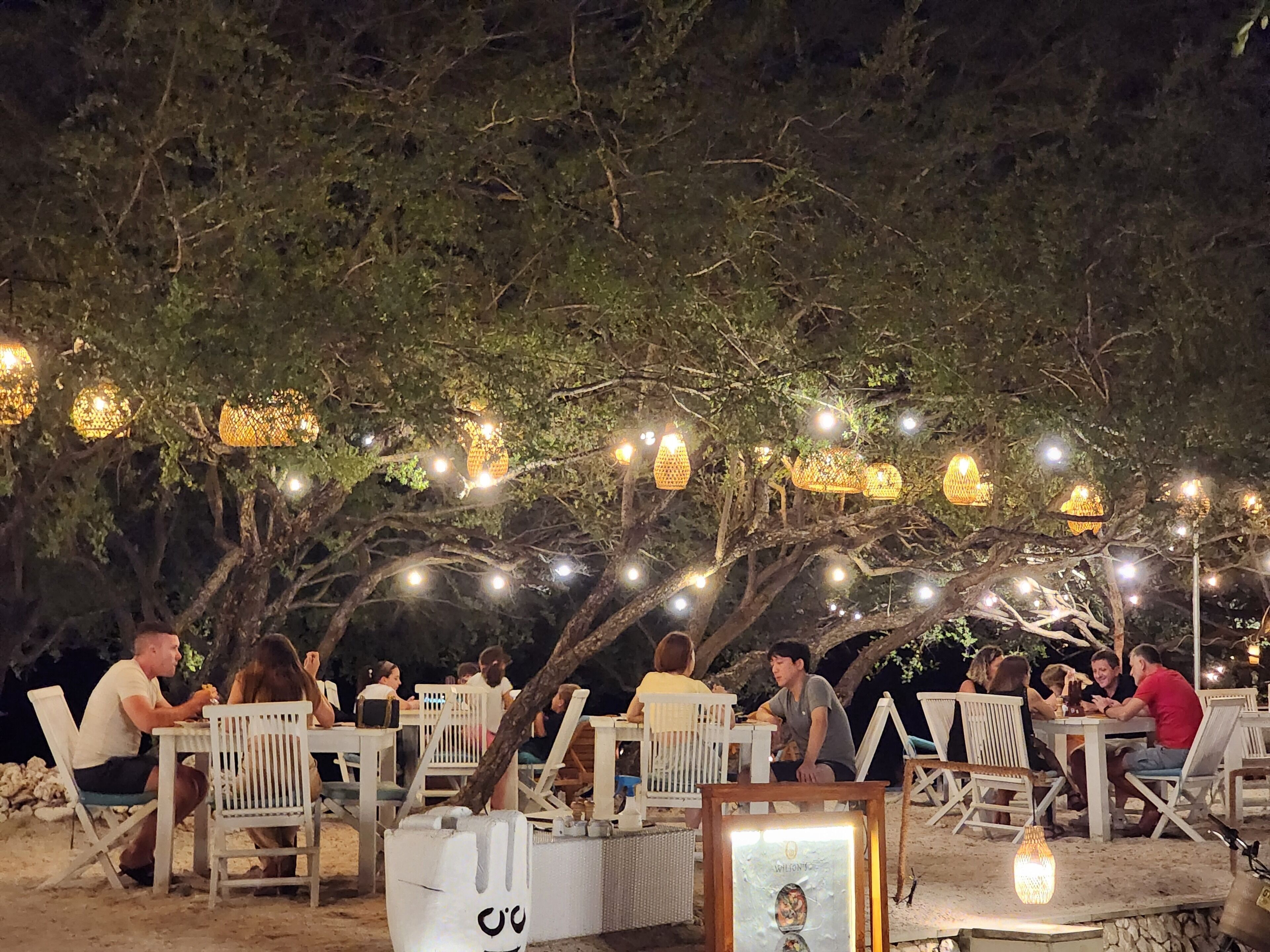 An outdoor dining area with white chairs and tables, string lights, and a framed picture on a table.