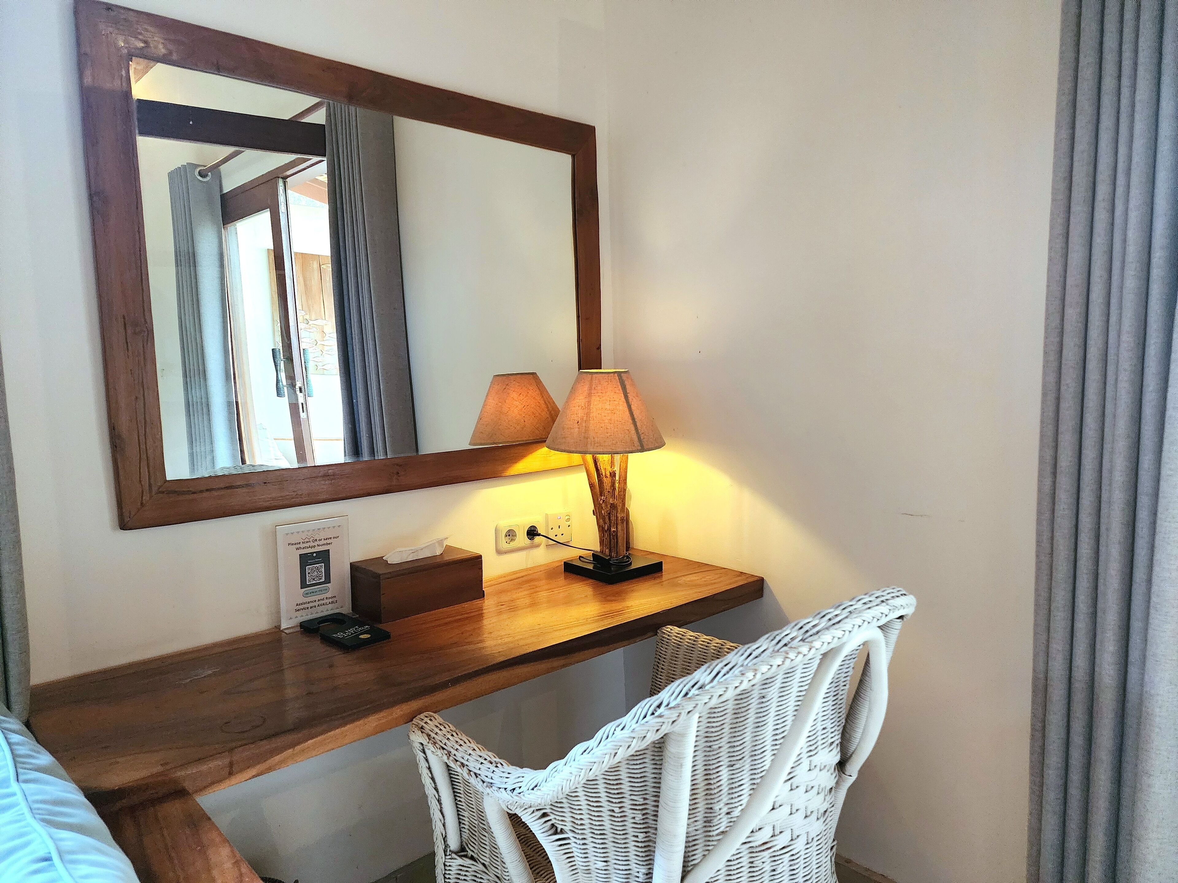 A wooden desk with a mirror, two lamps, a tissue box, and a wicker chair.