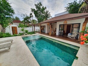 A swimming pool with a herringbone tile pattern, a covered patio area, and a view of a residential building with a white exterior and a red roof. - Wilsons Retreat (Gili Trawangan)