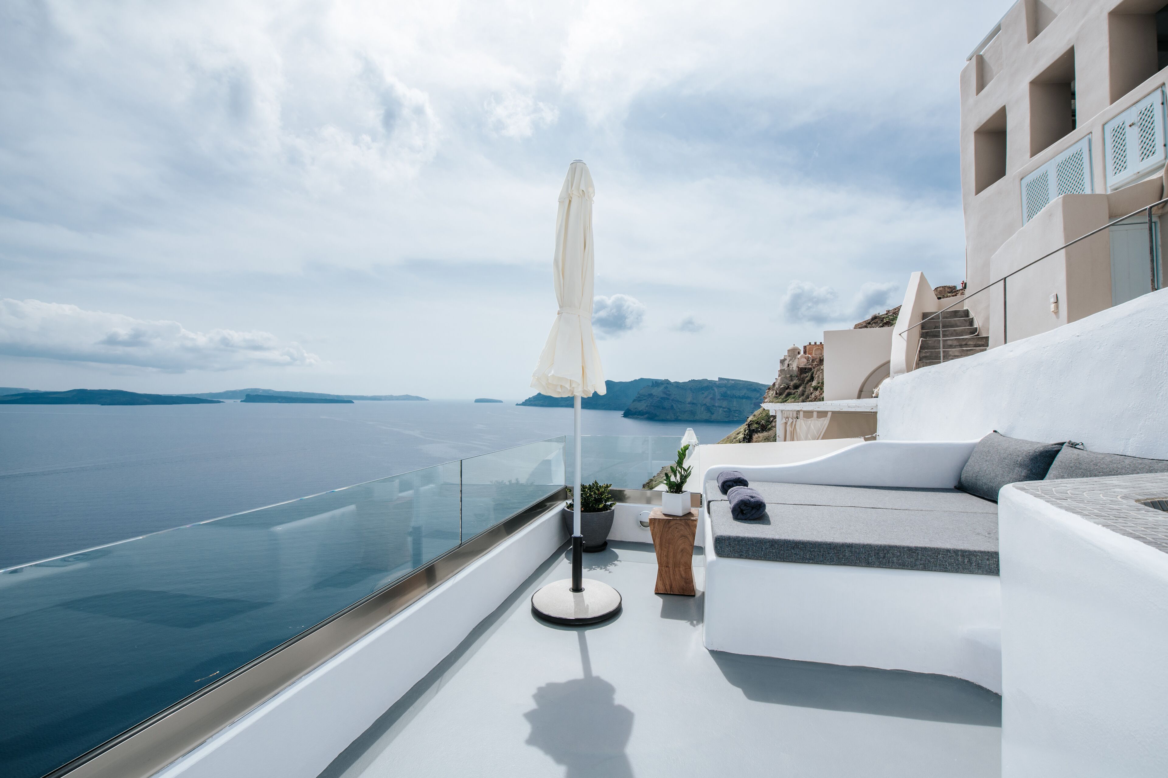 A modern balcony with a white sofa, a small table, and an umbrella. The balcony overlooks a body of water and a distant coastline.