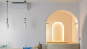 A modern interior with a white wall, a white ceiling, and a white sofa with patterned cushions. There is a white table with two stools, a book, and a bottle. An air conditioning unit is mounted on the wall. - Mykonos Moussa Suites (Mykonos)