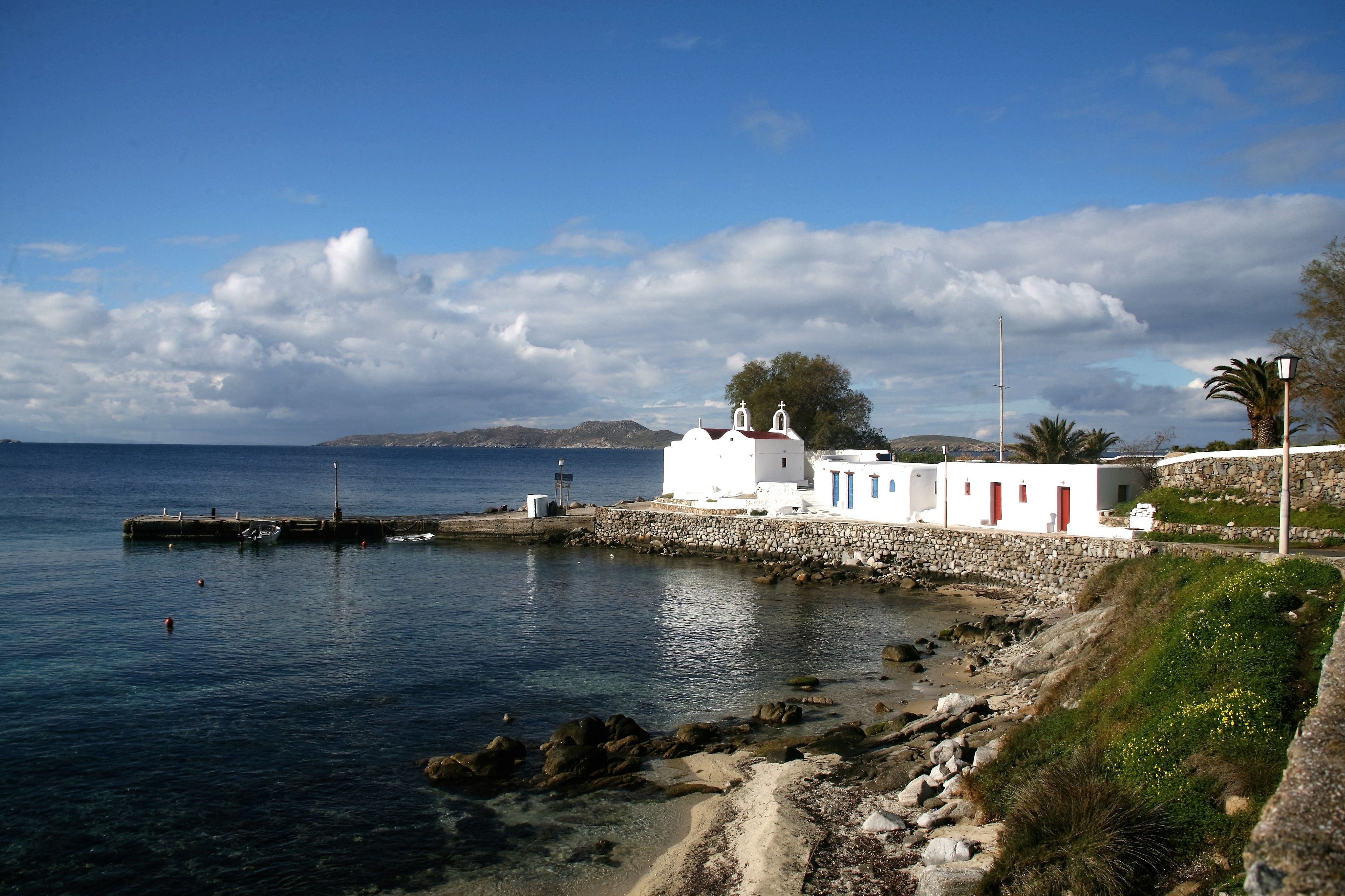 Escena costera con un edificio blanco con puertas rojas, un muelle de piedra y un cielo azul despejado.