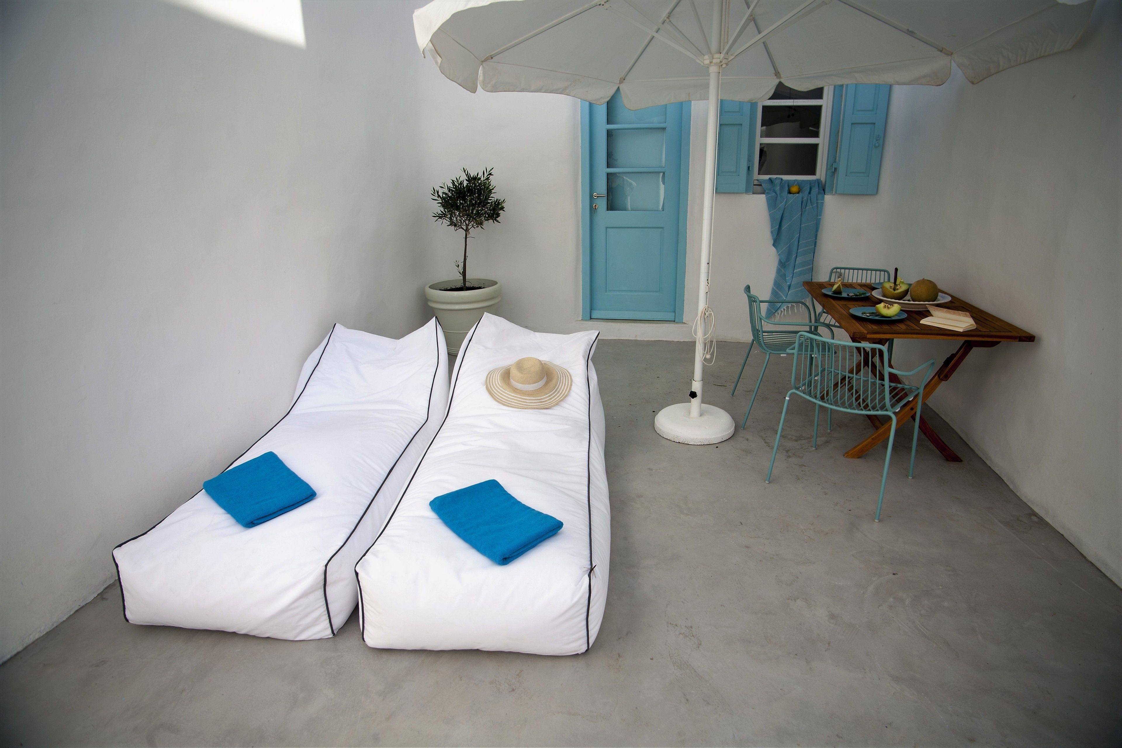 Una habitación con dos camas blancas, una mesita con una silla, una planta en maceta y una puerta azul.