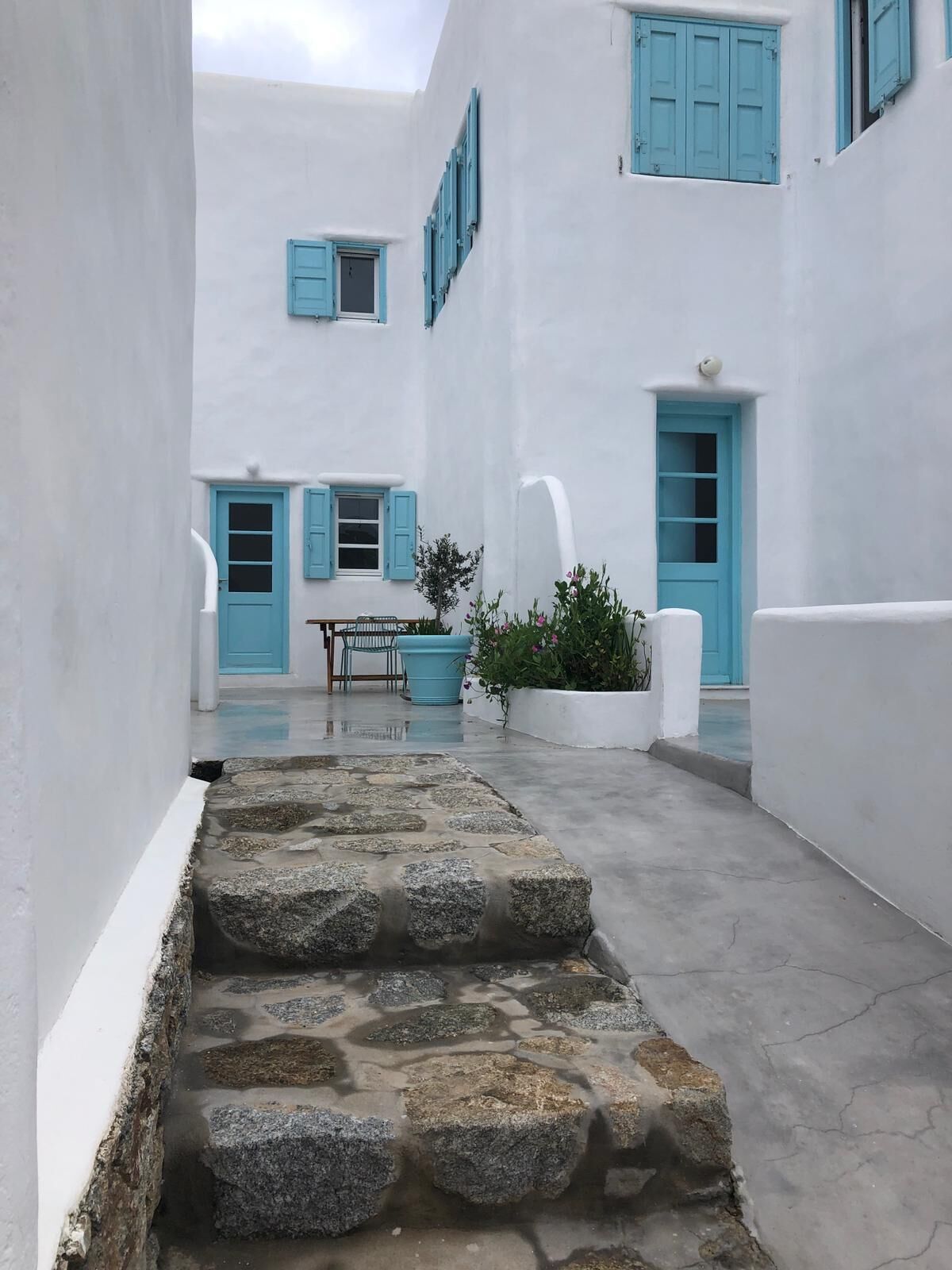 Un patio estrecho con paredes blancas y contraventanas azules, escalones de piedra que conducen a una puerta, y una pequeña mesa con sillas.