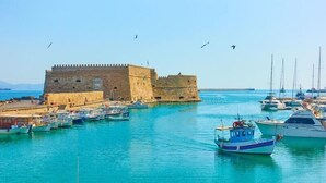 Un pueblo costero con una antigua fortaleza de piedra, un puerto deportivo con diferentes embarcaciones y aguas azul turquesa.