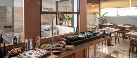 Montaje de bufé con distintos alimentos, una zona de cocina y un comedor con mesas y sillas.