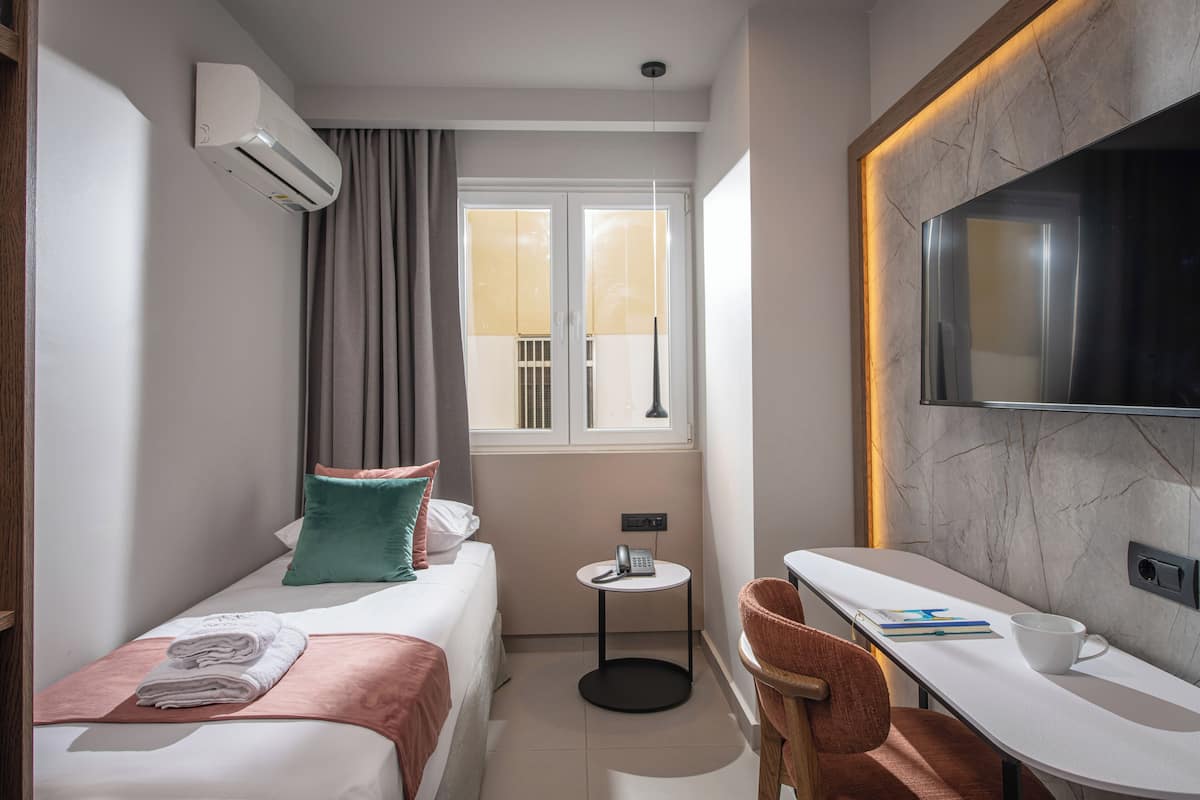Una habitación de hotel con cama, escritorio, silla, televisión, ventana con cortinas y aire acondicionado.