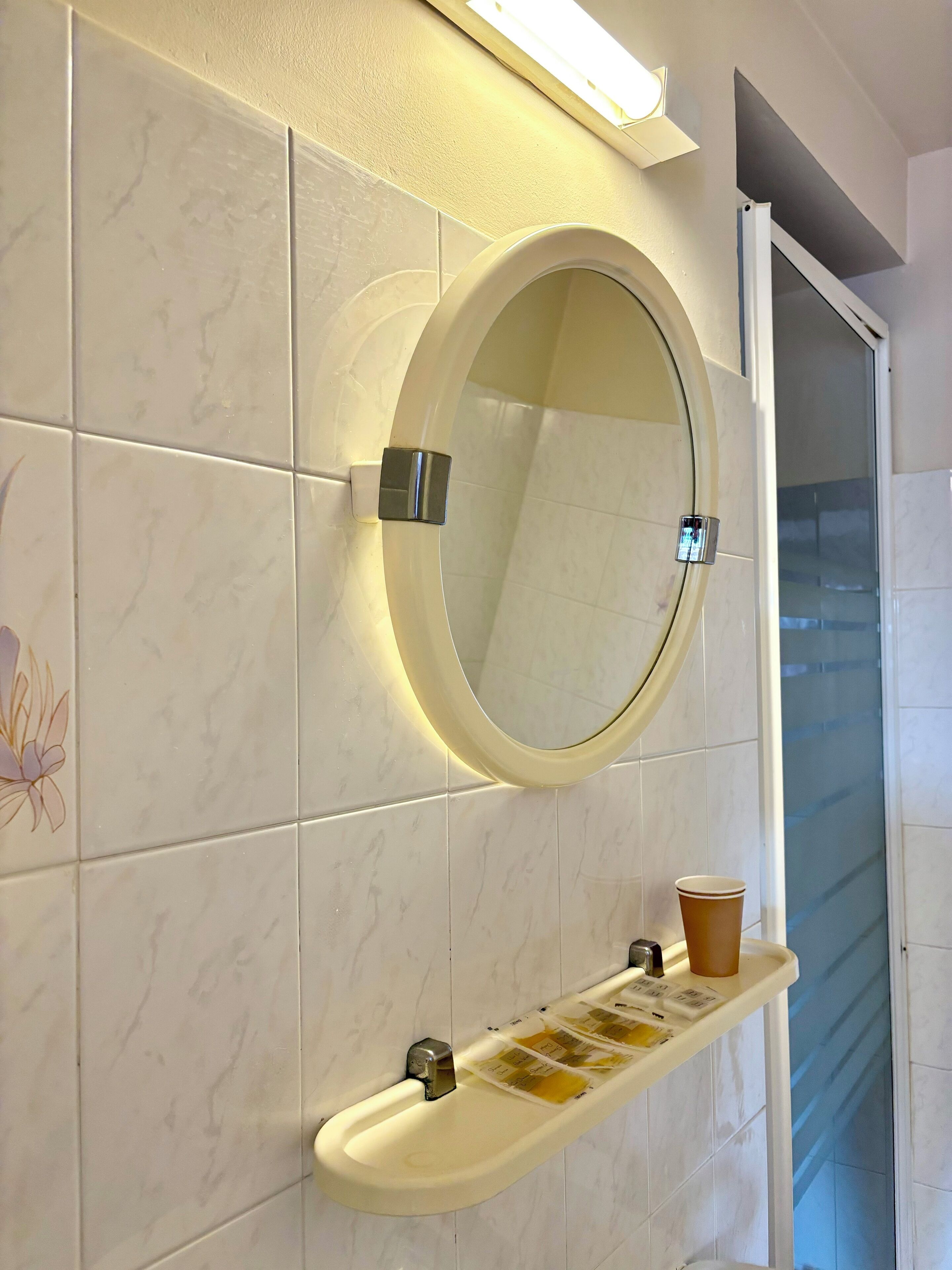 A bathroom with a round mirror, a shelf with toiletries, and a glass door leading to a shower area.