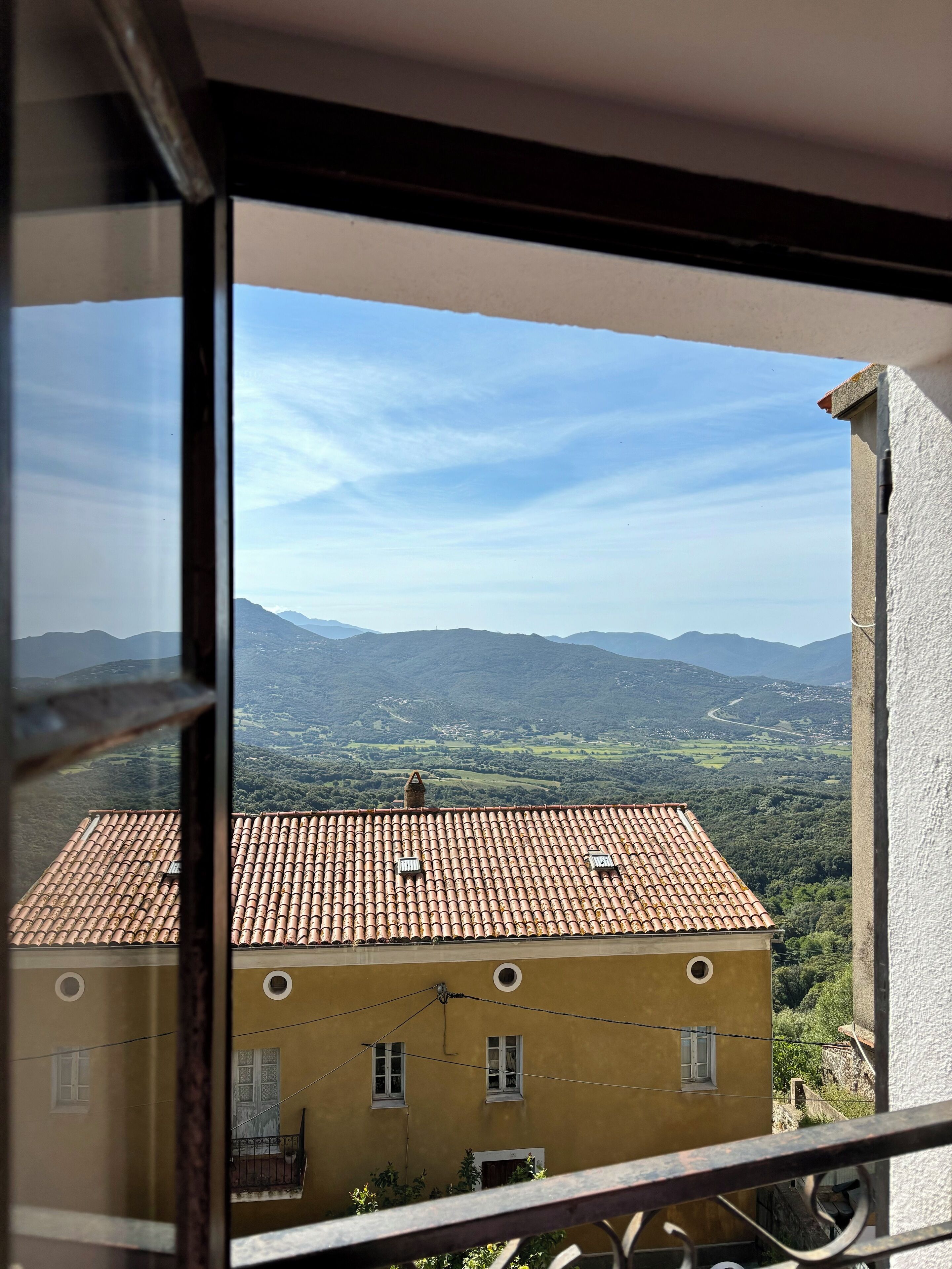 Vista dalla finestra su un edificio con tetto in terracotta e un balcone, con montagne e cielo sereno sullo sfondo.