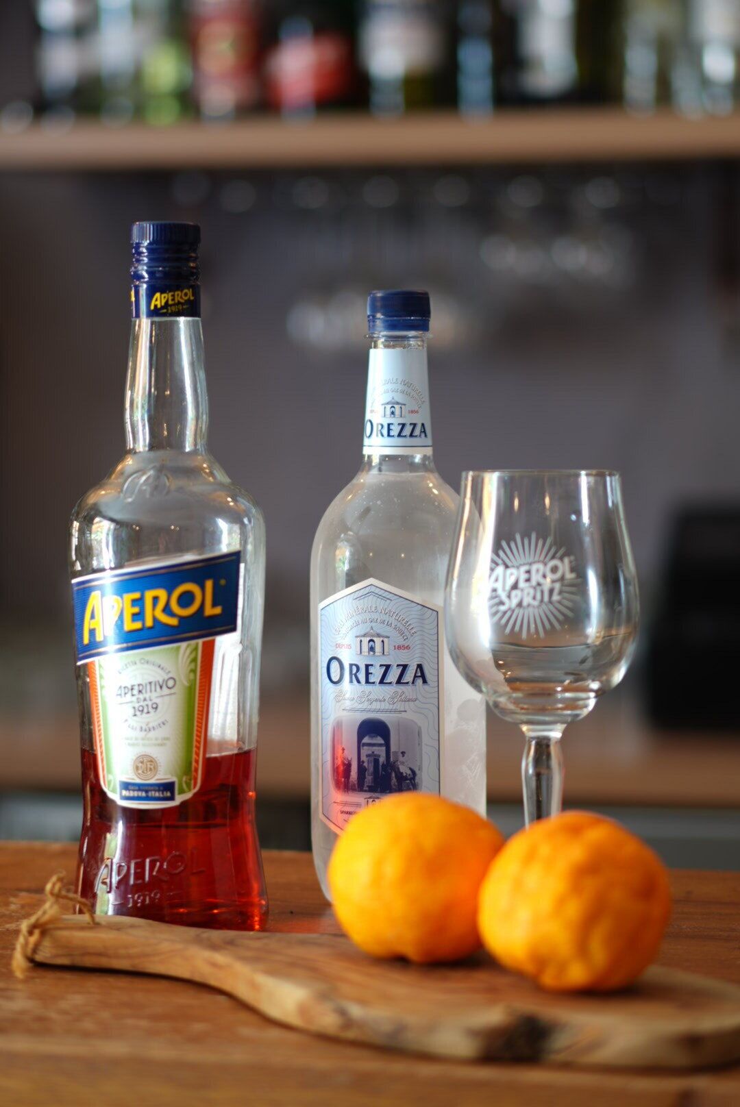 Deux bouteilles d’Aperol et d’OREZZA sur une surface en bois, avec deux oranges et un verre à vin.