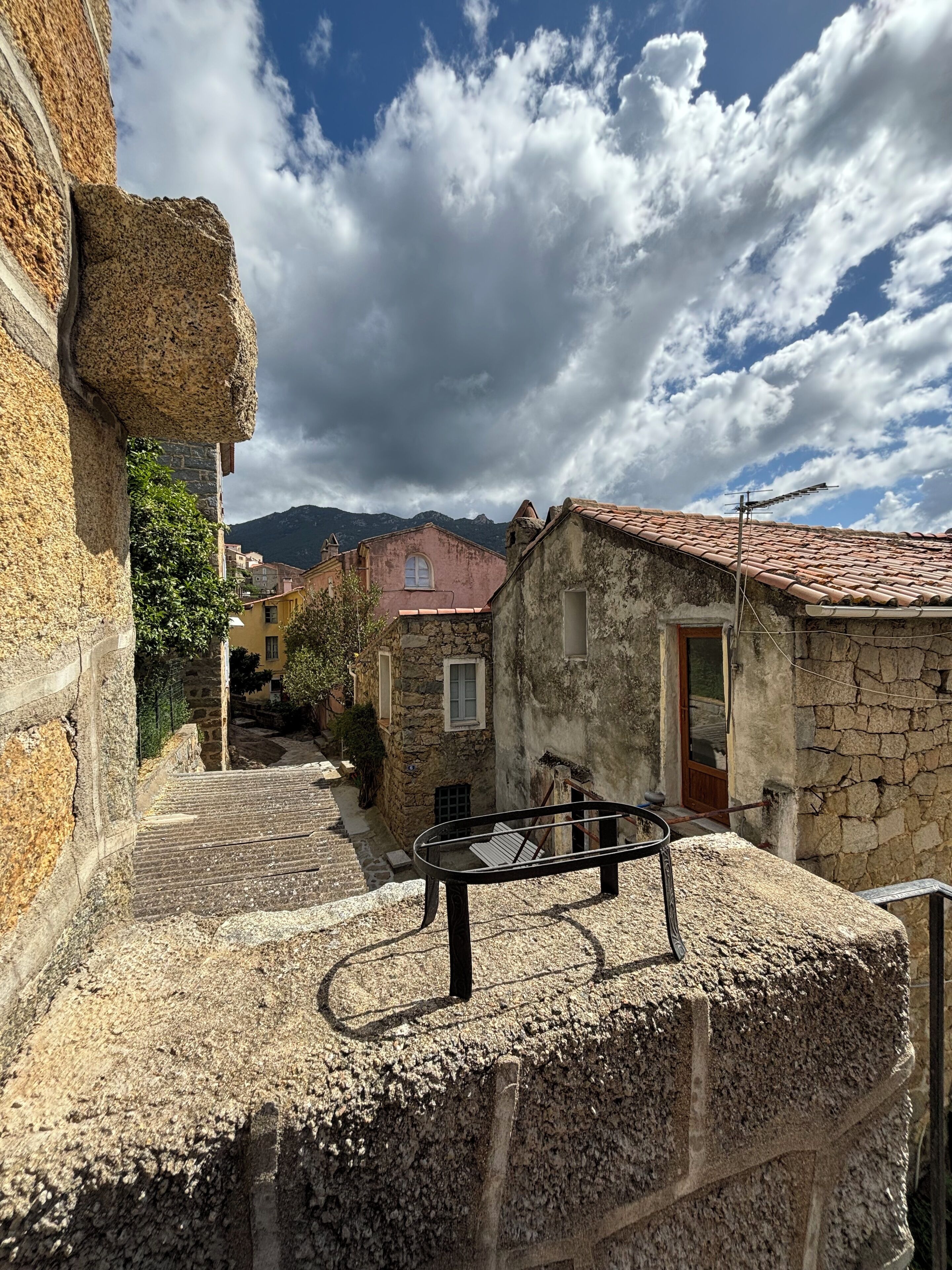 Una estrecha calle adoquinada con edificios de piedra, una barandilla de metal y una montaña al fondo.