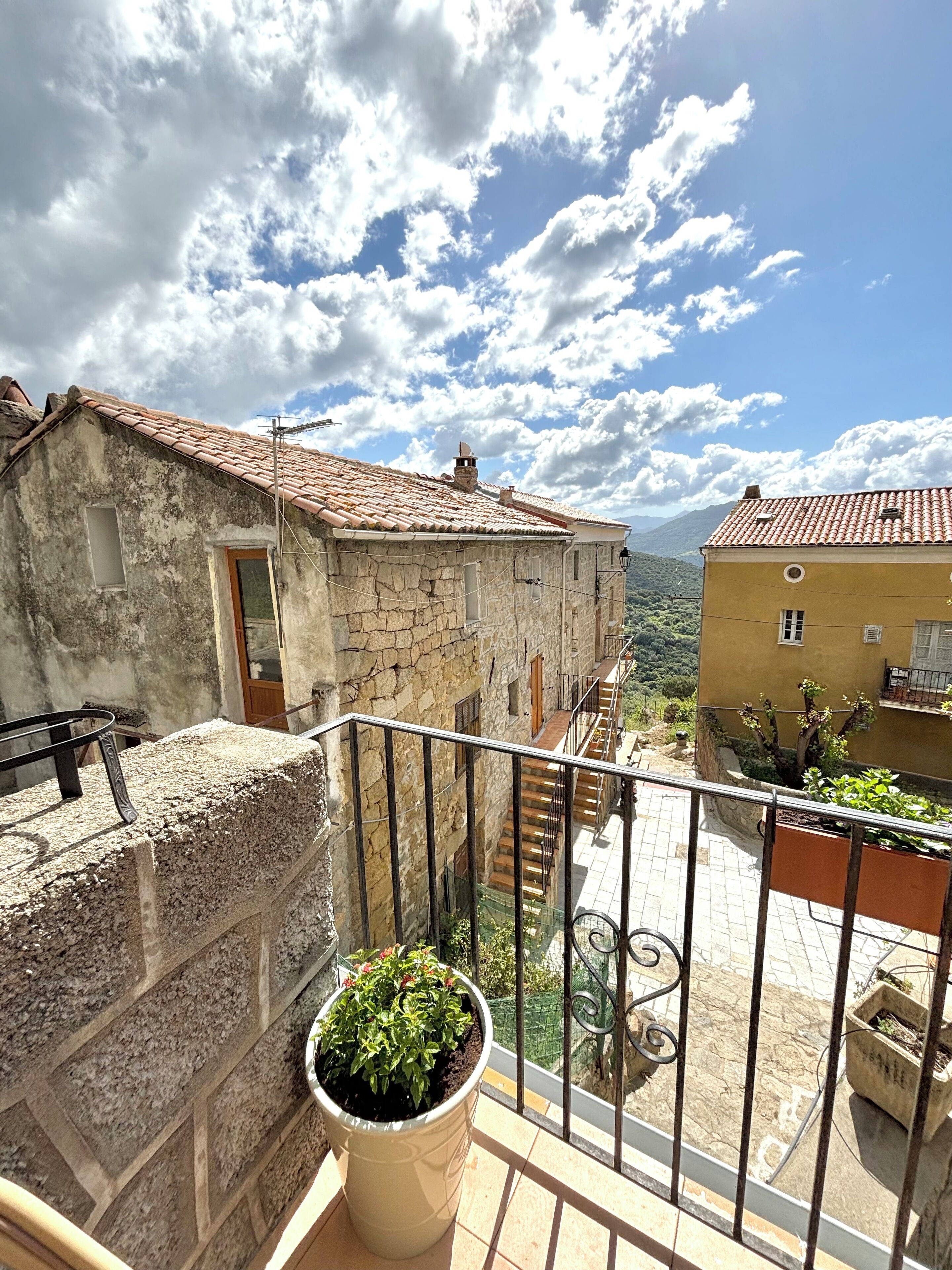 Un balcon agrémenté d’un pot de fleurs, des murs en pierre et une vue sur les bâtiments voisins et un paysage montagneux.