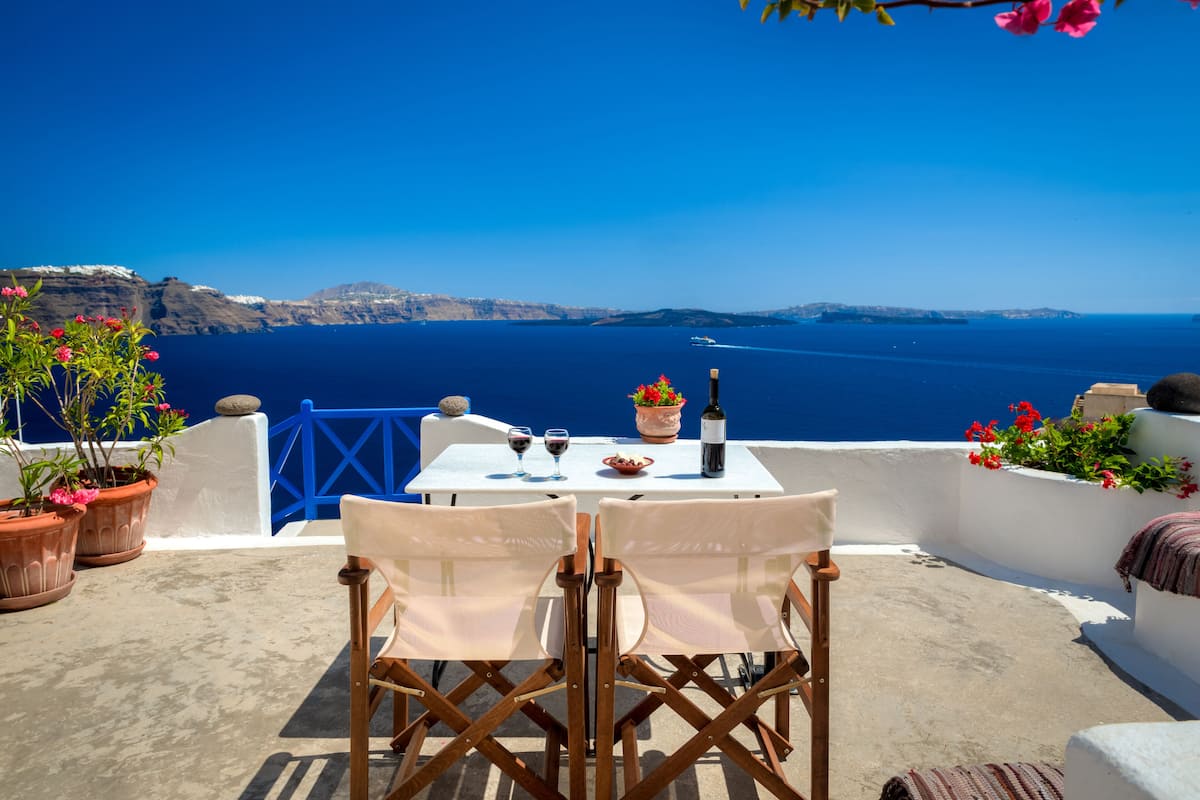 A table with two chairs on a terrace overlooking the sea, with potted plants and a bottle of wine.