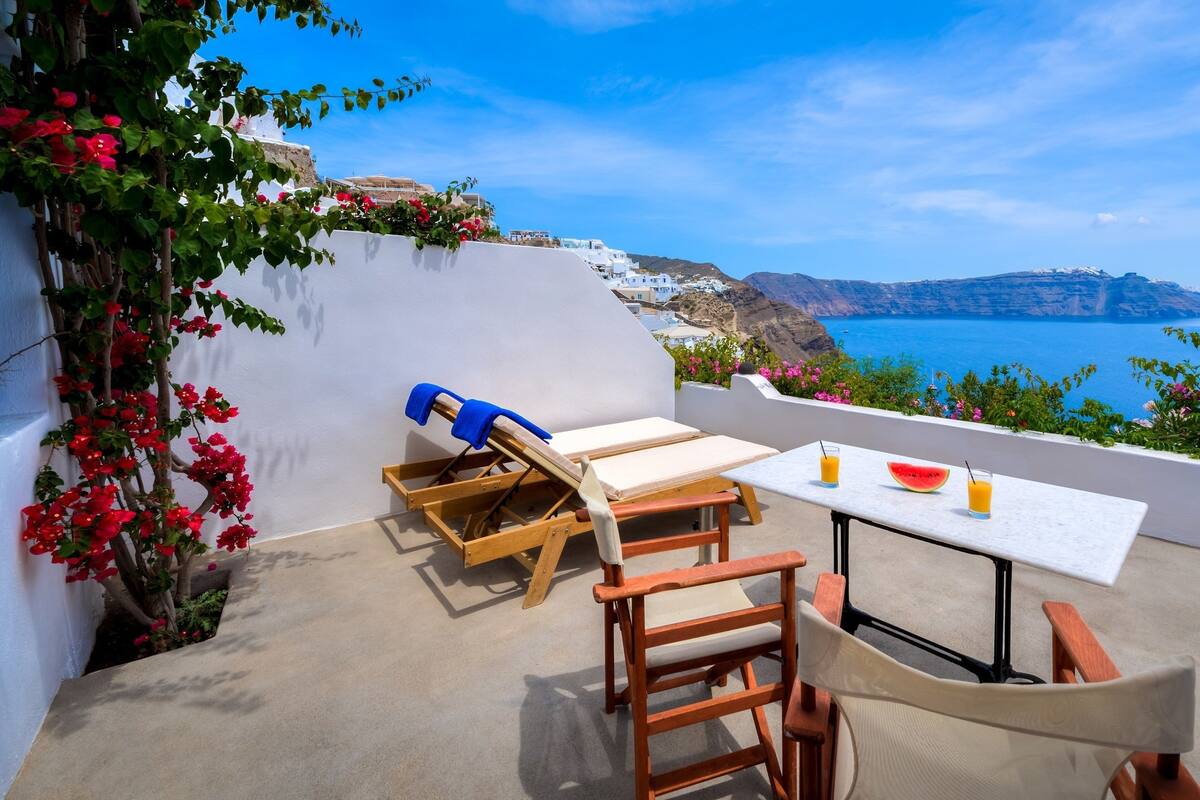 A terrace with a table, chairs, and a lounge chair overlooking a scenic coastal view.
