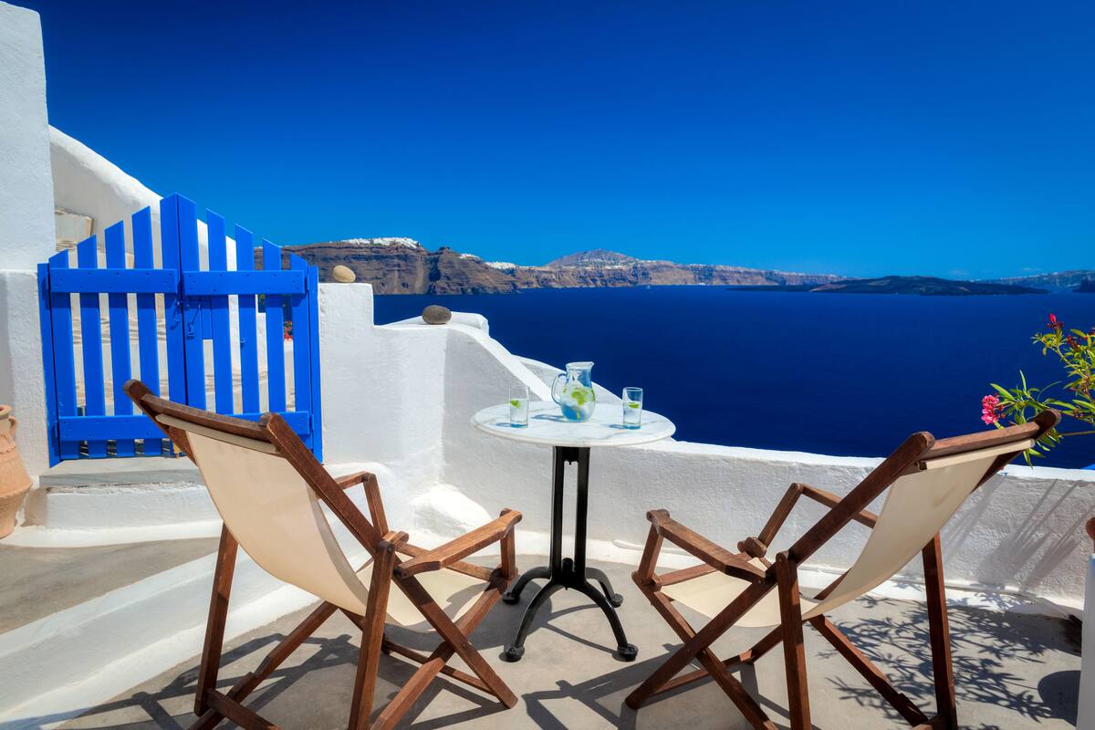 A white-walled terrace with a blue gate, two wooden chairs, a small round table, and a view of the sea and distant cliffs.