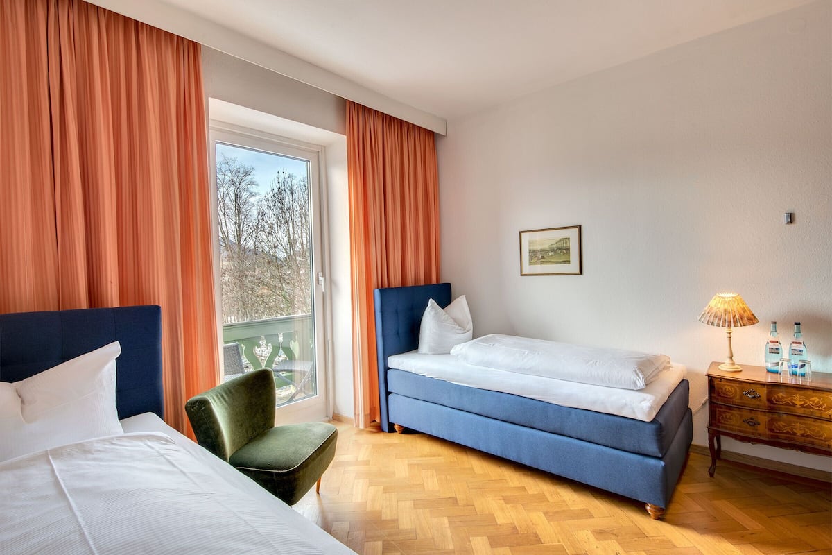 Ein Hotelzimmer mit zwei Betten, einem grünen Sessel, einer Holzkommode und einem Fenster mit Vorhängen.