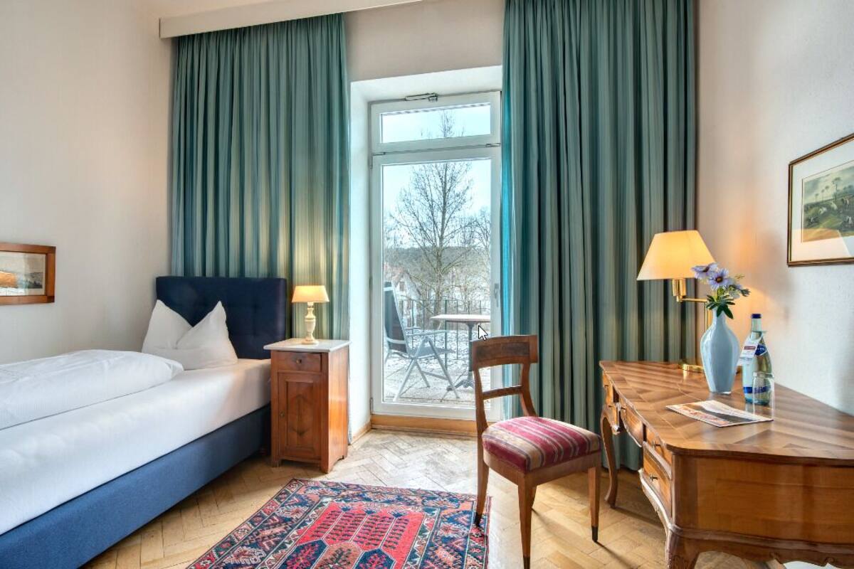Ein Hotelzimmer mit Bett, Schreibtisch, Stuhl, einem Fenster mit Vorhängen und Blick ins Freie.